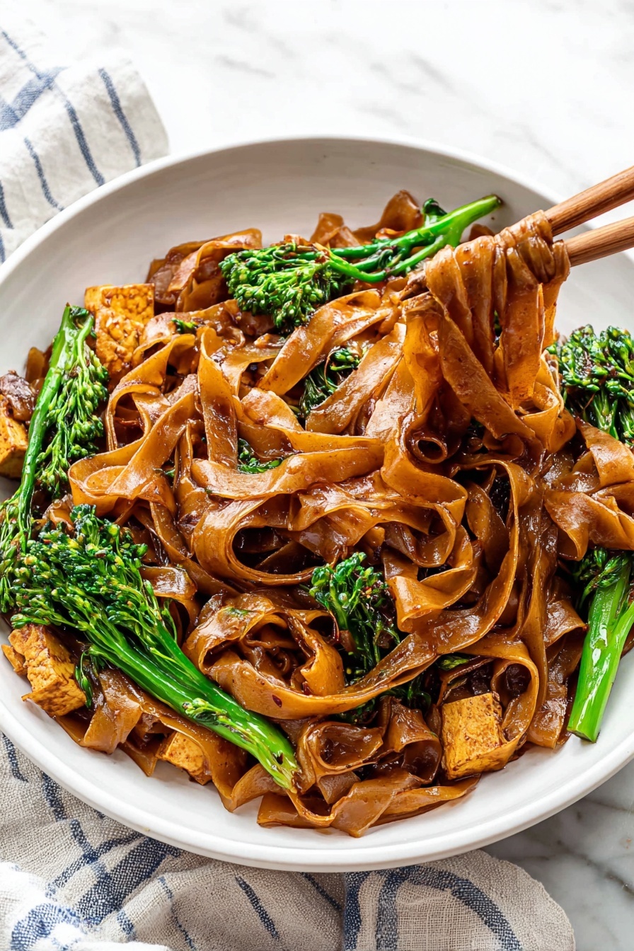 15 Minute Thai Pad See Ew Stir Fry, Thai stir fry recipes, quick Thai dinner ideas, authentic Pad See Ew, easy Thai noodle recipes - A white bowl is filled with wide, flat noodles coated in a shiny dark brown sauce, layered thick, with some noodles lifted by wooden chopsticks to the right side of the image. Mixed in are pieces of light brown cooked tofu and bright green broccolini with textured small florets. The bowl sits on a white cloth with blue stripes on a white marbled surface. The colors show a contrast between the dark noodles, green vegetables, and pale tofu, creating a rich and fresh look. photo taken with an iphone --ar 2:3 --v 7