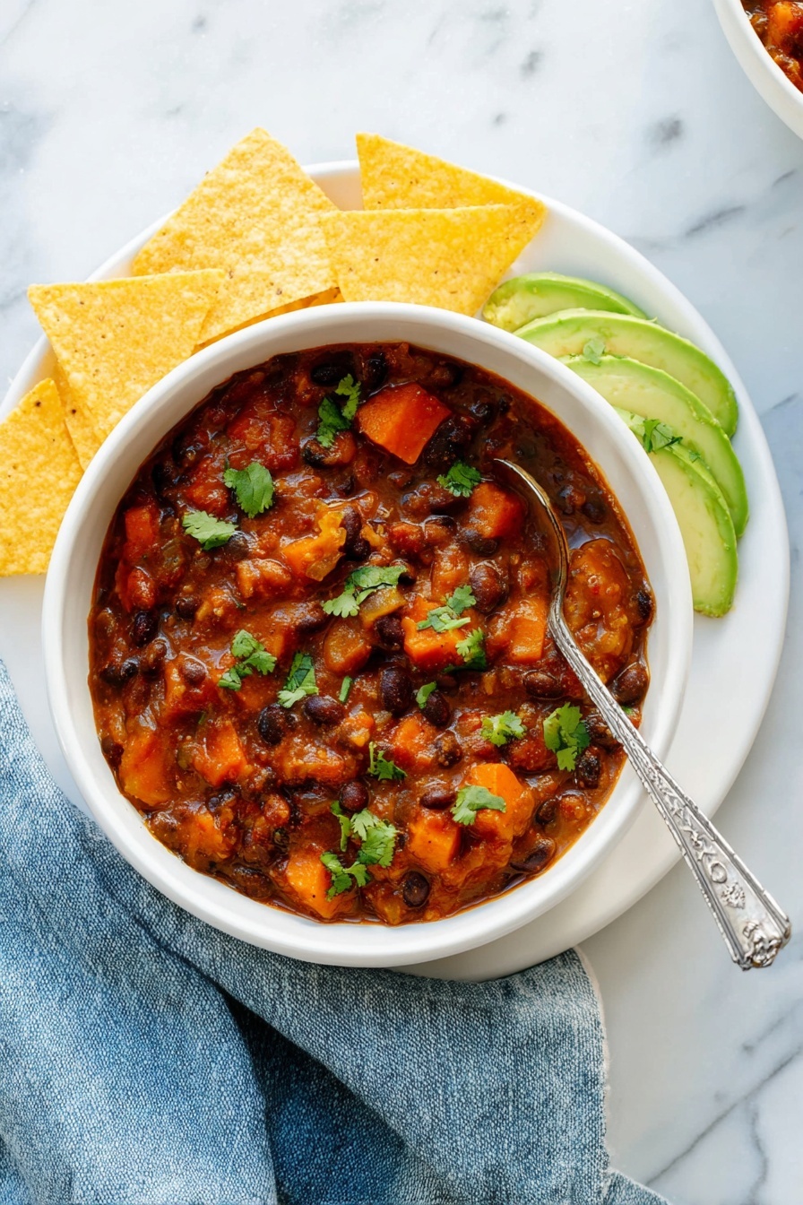 Vegetarian Chili, wholesome vegetarian chili, easy healthy chili, vegan chili recipe, hearty vegetable chili - A white bowl filled with thick chili containing black beans, orange carrot pieces, and chunks of sweet potato in a reddish-brown sauce, garnished with green cilantro leaves. On the right side inside the bowl are thin slices of light green avocado sprinkled with cilantro. The bowl sits on a white plate arranged with five triangular yellow tortilla chips around the left side. A silver spoon with a decorative handle rests inside the chili. The setting has a white marbled texture surface with a slightly crumpled blue cloth napkin next to the bowl. Photo taken with an iphone --ar 2:3 --v 7