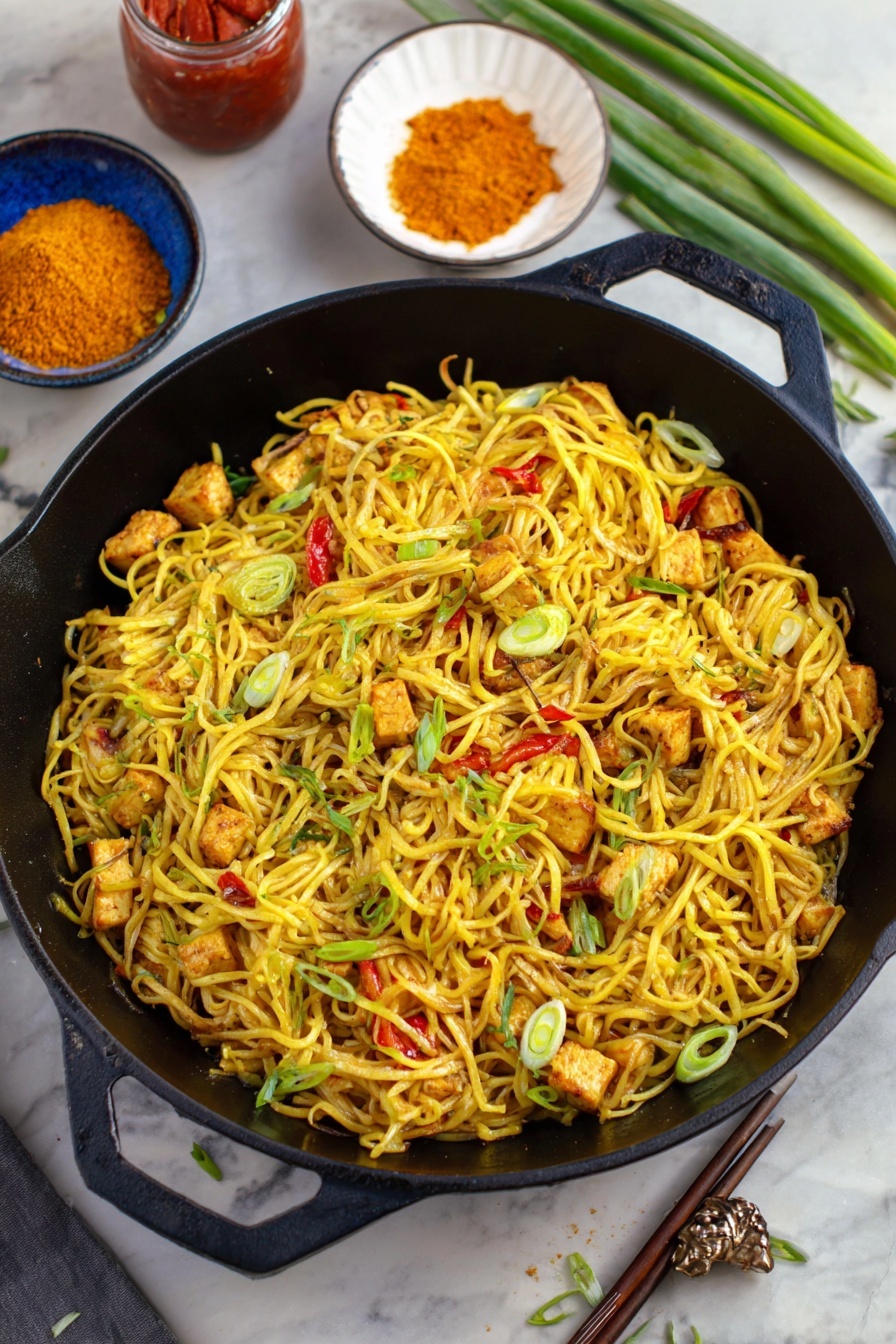 Easy Singapore Noodles, Singapore Noodles recipe, Asian noodle stir-fry, quick Singapore noodles, flavorful noodle dishes - A black cast iron pan filled with several layers of yellow noodles mixed with small pieces of grilled tofu that show a light brown char, scattered with thin slices of green onions and small bits of red bell pepper. The noodles have a slight shine and look seasoned. Around the pan, there is a white bowl with orange powder, a jar with red sauce, a blue bowl with chopsticks placed on the side, and some green onion stalks on a white marbled surface. The scene has a mix of greens and yellows that make the dish look fresh and tasty photo taken with an iphone --ar 2:3 --v 7