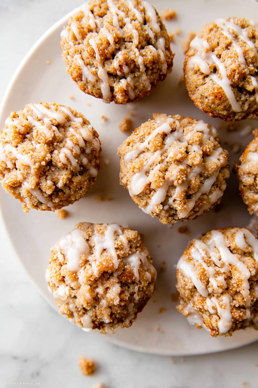 Pumpkin Crumb Muffins, Fall Pumpkin Muffins, Pumpkin Muffin Recipe, Easy Pumpkin Muffins, Cinnamon Pumpkin Muffins - The image shows a white plate with six small muffins arranged in a loose circle on a white marbled surface. Each muffin has a crumbly, light brown streusel topping that covers the whole top and looks crunchy. A thin layer of white icing is drizzled unevenly over the muffins, creating shiny lines that drip slightly onto the plate. A few crumbs are scattered around the plate and on the marbled surface. The muffins look moist and textured with a mix of crumbly and smooth parts. Photo taken with an iphone --ar 2:3 --v 7