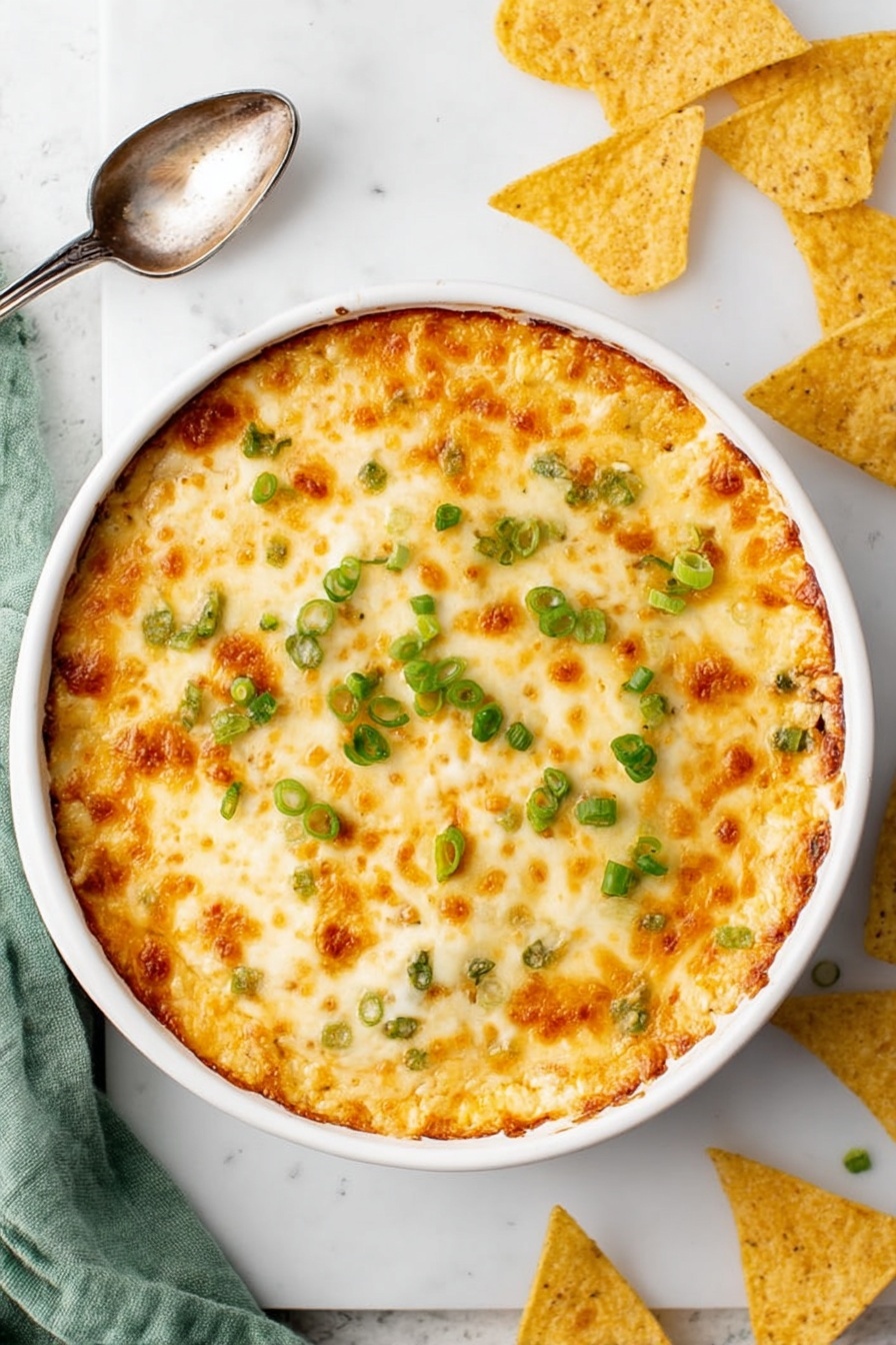 Crab Rangoon Dip with Wonton Chips, crab dip appetizers, creamy crab dip recipe, easy wonton chip recipe, crowd-pleasing game day dip - A round white baking dish filled with a golden, bubbly cheese layer on top, showing patches of light brown where the cheese is slightly toasted, scattered with small rings of bright green chopped scallions. The entire dish sits on a white marbled surface with a vintage silver spoon to the side, broken and whole triangular yellow tortilla chips nearby, and a soft green cloth partly visible at the edge. Photo taken with an iphone --ar 2:3 --v 7