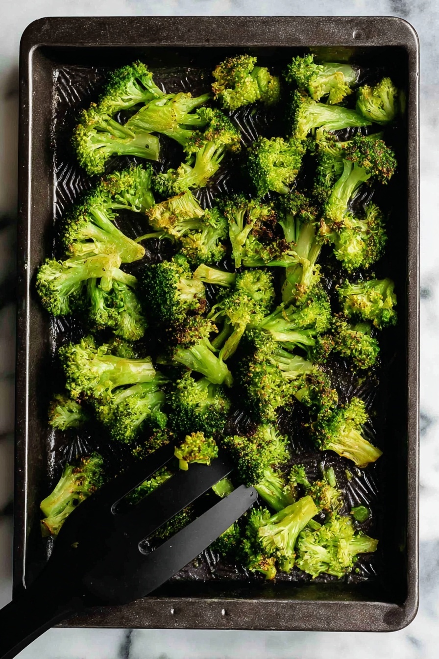Oven Roasted Broccoli with Garlic and Paprika, roasted broccoli recipes, healthy roasted broccoli, garlic broccoli side dish, smoky broccoli roasted - A dark rectangular baking tray holds a single layer of cooked broccoli florets. The broccoli pieces are bright green with some slightly browned tips, showing a crispy texture. The florets vary in size and shape, spread unevenly across the tray. In the bottom right corner, a black spatula is lifting some broccoli. The tray rests on a white marbled surface. photo taken with an iphone --ar 2:3 --v 7