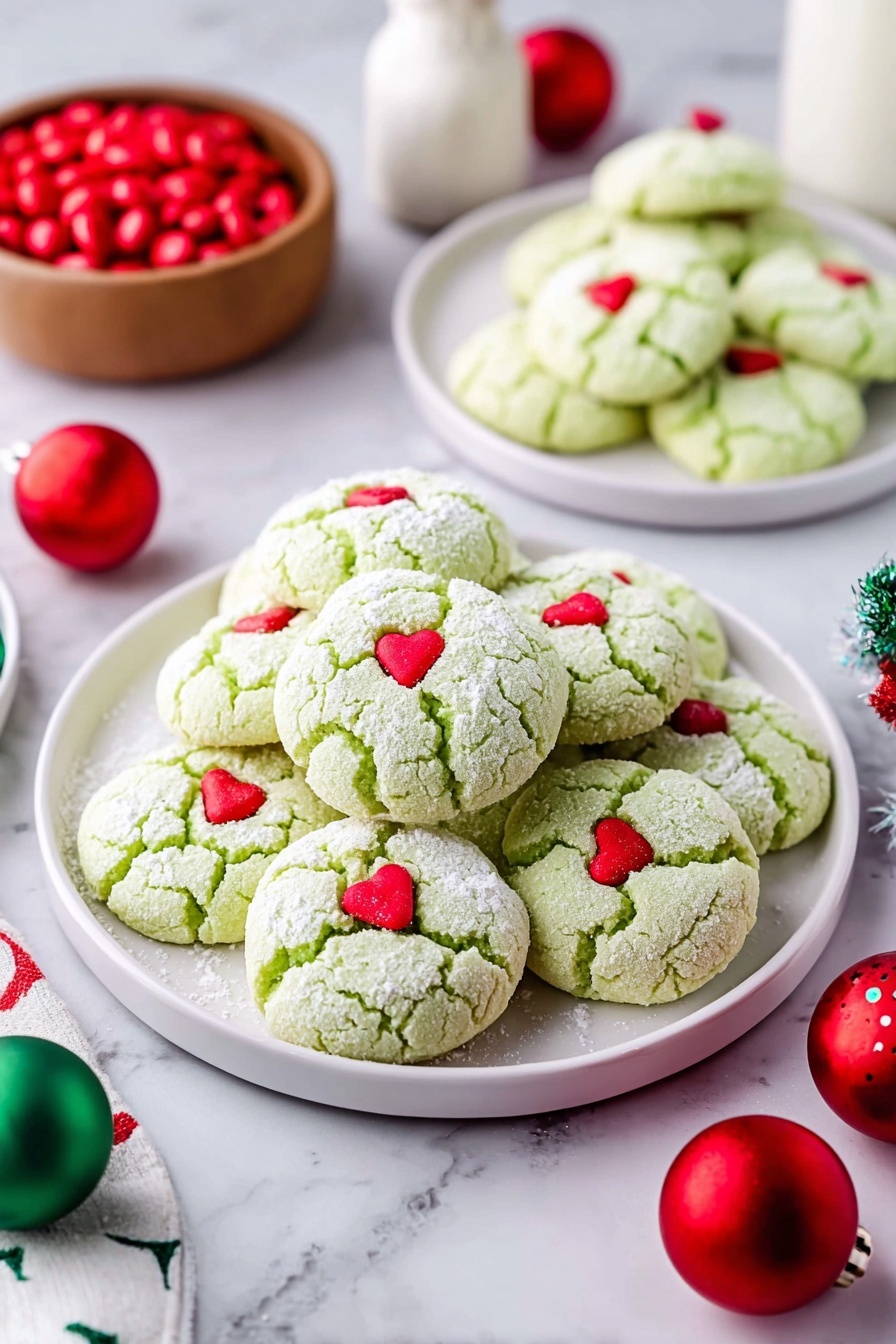 Green Peppermint Cake Mix Cookies, peppermint cake mix cookies, holiday peppermint cookies, easy peppermint cookie recipe, festive green cookies - A large pile of light green cookies with a soft cracked texture, each topped with a small red heart candy, sits on a white plate in the center. Two smaller white plates in the background hold more of the same cookies. The cookies are dusted lightly with powdered sugar. Around the plates are red and green round Christmas ornaments and a small wooden bowl filled with red candies on a white marbled surface. A white bottle is visible in the background. Photo taken with an iphone --ar 2:3 --v 7