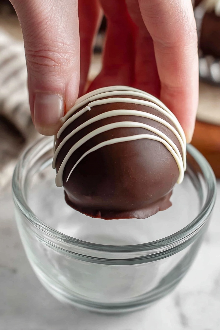 Homemade Hot Chocolate Bombs, hot chocolate bombs, how to make hot chocolate bombs, chocolate sphere hot chocolate, festive hot chocolate drinks - A woman's hand is holding a round dark chocolate ball with white thin lines of icing drizzled vertically on top. The chocolate ball has two layers, the outer smooth dark chocolate shell, and the white icing on the top. It is being held above a clear glass cup with a simple design. The background shows a white marbled textured surface. photo taken with an iphone --ar 2:3 --v 7
