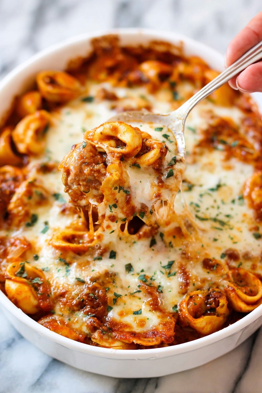 Cheesy Baked Tortellini with Beef, baked tortellini with beef, cheesy pasta bake, hearty meat tortellini casserole, comforting baked tortellini recipe - A white dish filled with baked tortellini pasta tossed in a rich red tomato sauce and covered with a thick layer of melted, golden-brown cheese with green herb sprinkles on top. Some tortellini are visible under the cheese, showing their smooth curved shapes and orange sauce coating. A woman's hand holds a silver spoon lifting a portion of the cheesy pasta, with melted cheese stretching slightly. The background shows more pasta inside the white dish on a white marbled surface. photo taken with an iphone --ar 2:3 --v 7