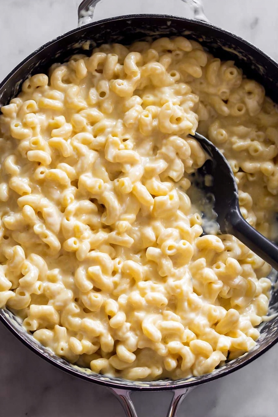 Cheddar Baked Mac and Cheese, baked mac and cheese recipe, cheesy baked pasta, easy mac and cheese casserole, comfort food recipes - The image shows a black pan filled with creamy macaroni and cheese. There is one thick layer of soft elbow pasta coated in smooth, light yellow cheese sauce. A black spoon is partly submerged in the macaroni, lifting a scoop on one side of the pan. The pan is set on a white marbled surface which is slightly visible around the edges. Photo taken with an iphone --ar 2:3 --v 7