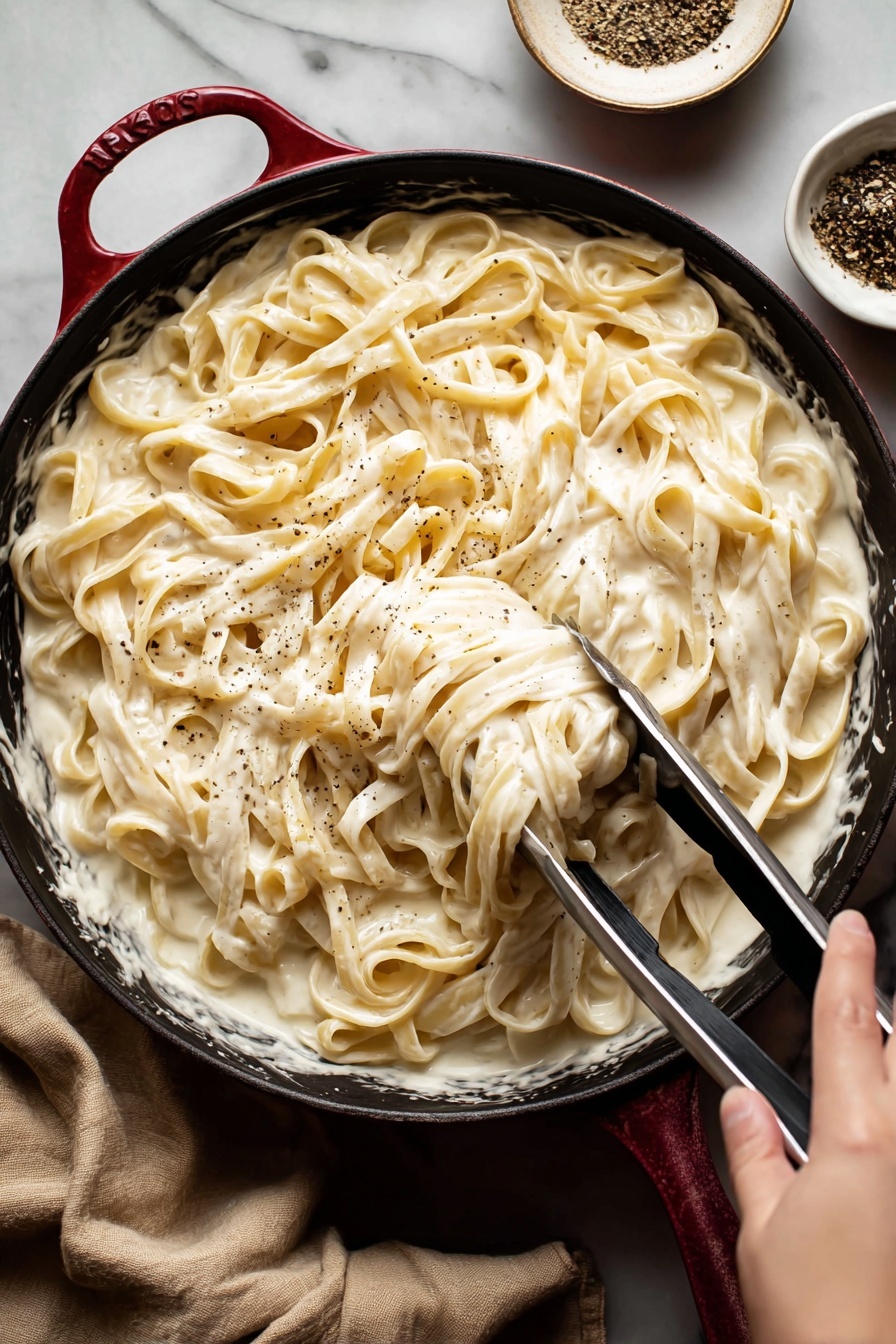 Vegan Cashew Alfredo Sauce, vegan Alfredo sauce, plant-based Alfredo, dairy-free Alfredo sauce, gluten-free Alfredo sauce - A close-up view of creamy fettuccine pasta served in a black pan with a red handle edge, the pasta is covered in a thick, smooth white sauce that looks rich and velvety; the long pasta strands are mixed well with the sauce and lightly sprinkled with black pepper; a woman's hand is holding metal tongs, lifting some pasta from the pan; in the background, there is a small white bowl with black pepper, and the whole setting is on a white marbled surface with a beige cloth nearby photo taken with an iphone --ar 2:3 --v 7