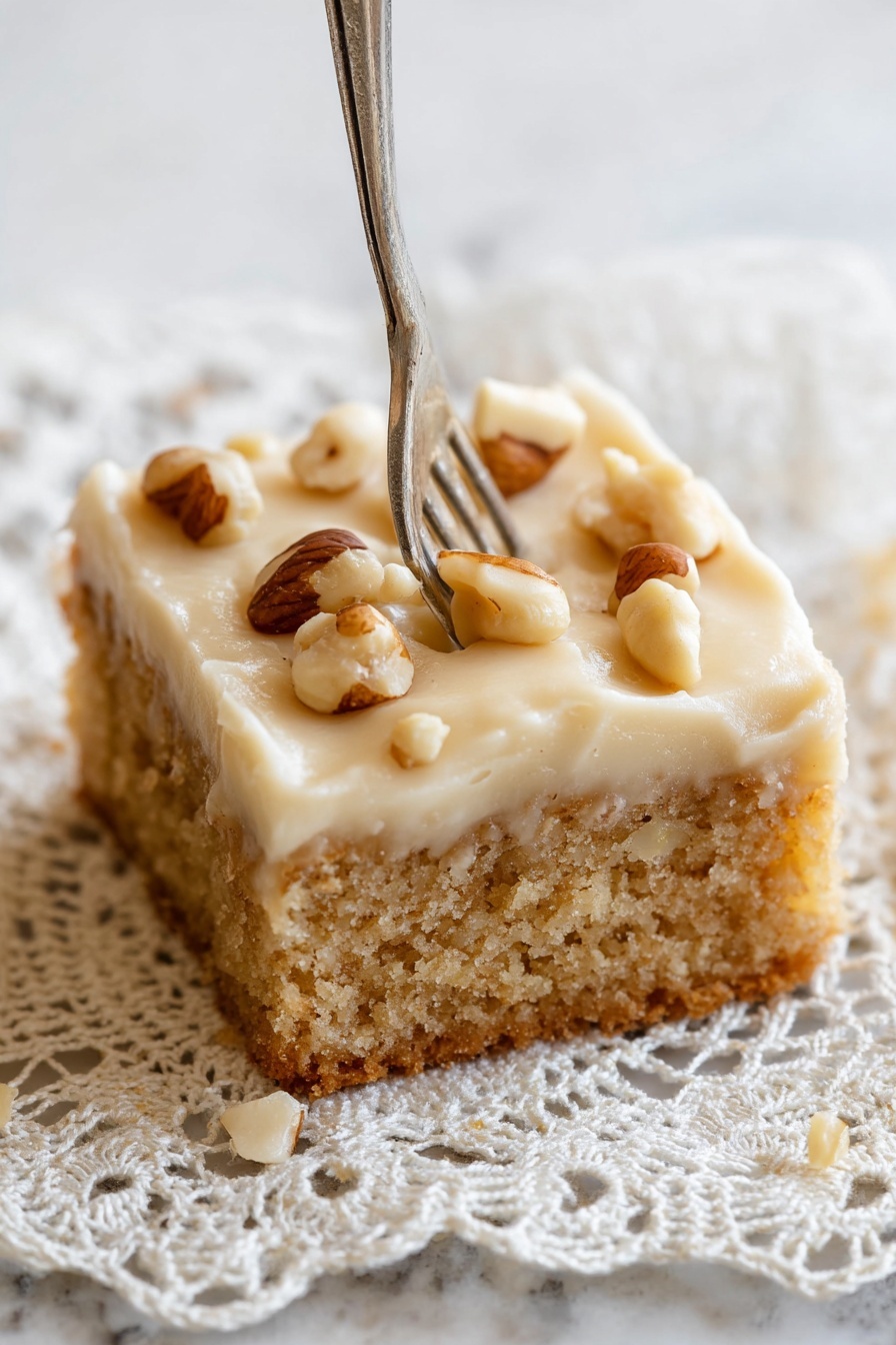 Peanut Butter Cake with Creamy Frosting, peanut butter cake recipe, easy peanut butter cake, moist peanut butter cake, nutty dessert ideas - A close-up of a single square piece of cake with two layers, the bottom layer is a light brown, soft, crumbly cake base, and the top layer is a thick, pale cream frosting with a smooth texture. The frosting is decorated with whole, shiny brown and white nuts scattered unevenly on top. A fork is pressing slightly into the right side of the frosting. The cake rests on a delicate crochet lace doily with intricate patterns, placed on a white marbled surface. Photo taken with an iphone --ar 2:3 --v 7