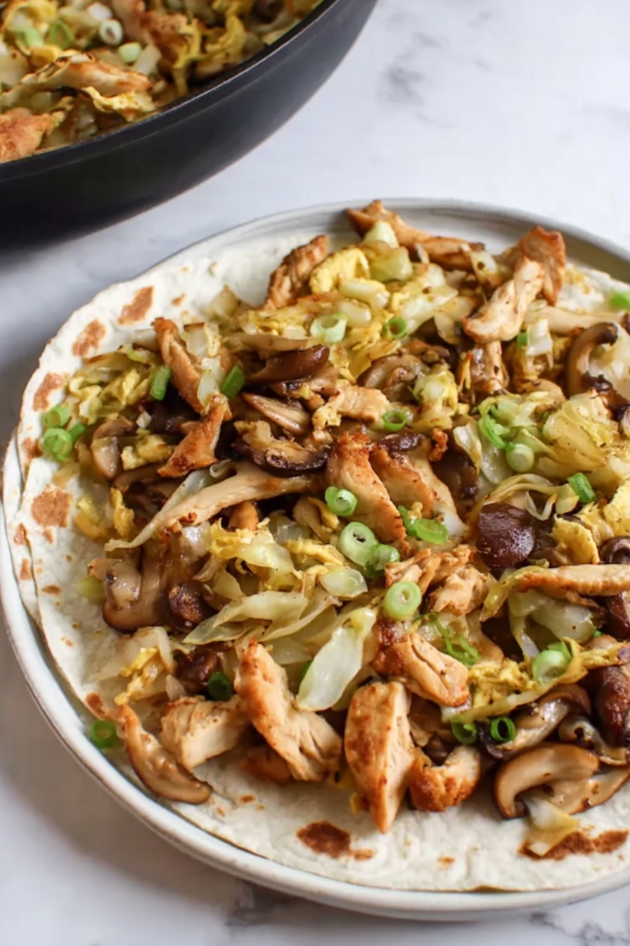 Moo Shu Chicken, Chinese takeout style Moo Shu Chicken, easy Chinese stir-fry, quick weeknight dinner, homemade Moo Shu Chicken - This image shows a close-up of a black frying pan filled with a stir-fry dish. The dish has many pieces of brown, cooked chicken strips mixed with light yellow cabbage slices and orange-yellow egg strips. There are also some sliced mushrooms with a dark brown outer edge and light beige inside, scattered throughout. Small green onion pieces are sprinkled over the dish adding a splash of green color. The frying pan is set on a white marbled surface. photo taken with an iphone --ar 2:3 --v 7