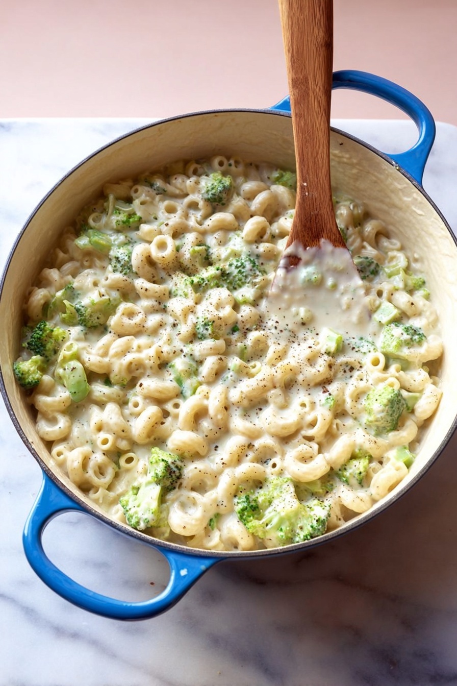 Creamy Broccoli Pasta, creamy broccoli pasta recipe, easy broccoli pasta, vegan broccoli pasta, healthy pasta with broccoli - A large white pot with blue handles is filled with small tube pasta and small green broccoli pieces mixed in a thick, creamy white sauce with visible black pepper specks, all stirred by a wooden spoon standing upright in the pot, placed on a white marbled surface with a soft pink background, photo taken with an iphone --ar 2:3 --v 7