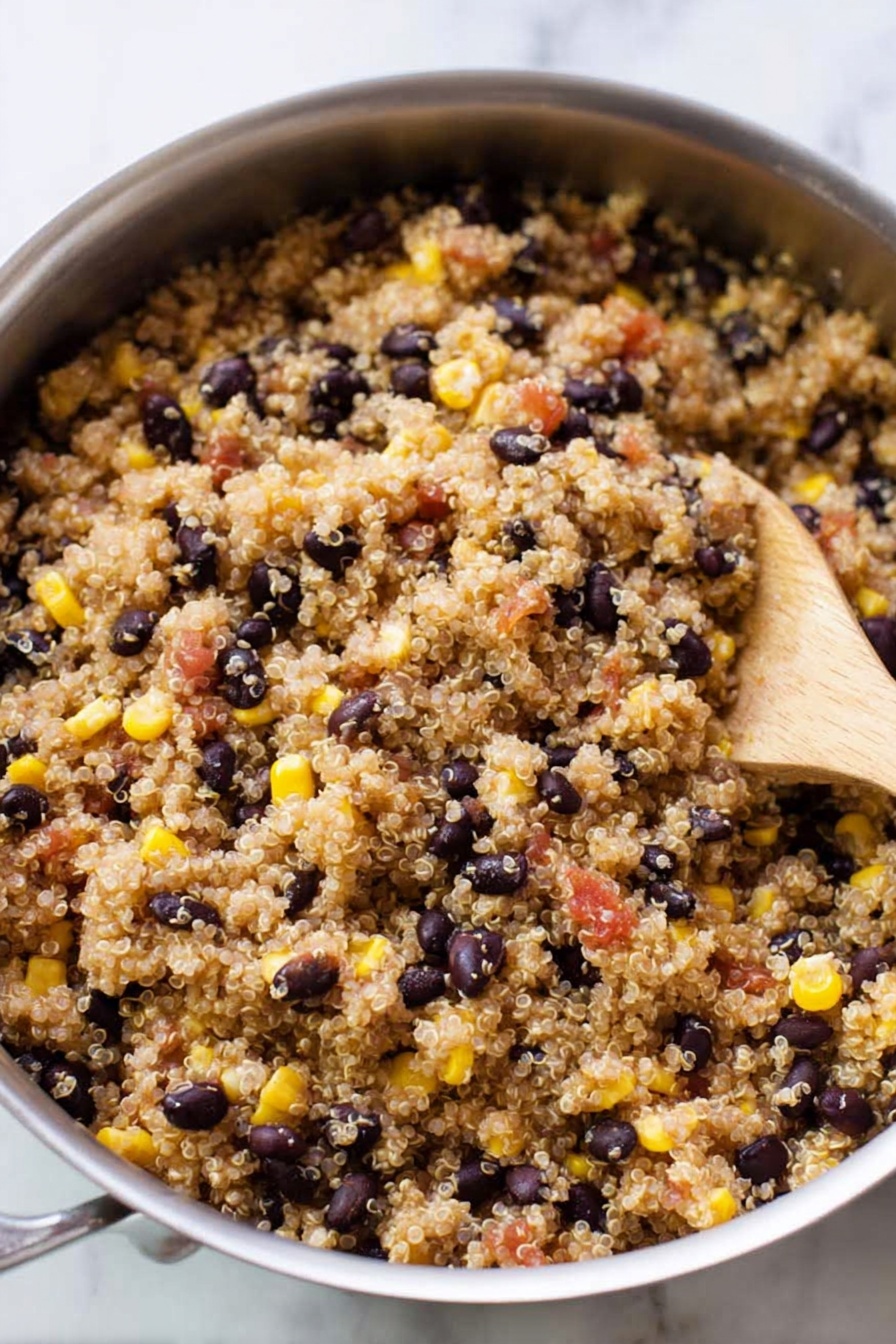 Easy Mexican Quinoa Bowl, Mexican quinoa bowl, healthy quinoa bowl, quick vegetarian dinner, flavorful quinoa salad - A silver pan filled with cooked quinoa mixed with black beans, yellow corn, and small pieces of red tomatoes. The quinoa grains are light brown and fluffy, evenly spread throughout the pan. The black beans are scattered, showing their dark smooth texture while the yellow corn adds bright pops of color. A wooden spoon rests inside the pan on the right side, partially covered by the quinoa mixture. The background is a white marbled surface. photo taken with an iphone --ar 2:3 --v 7