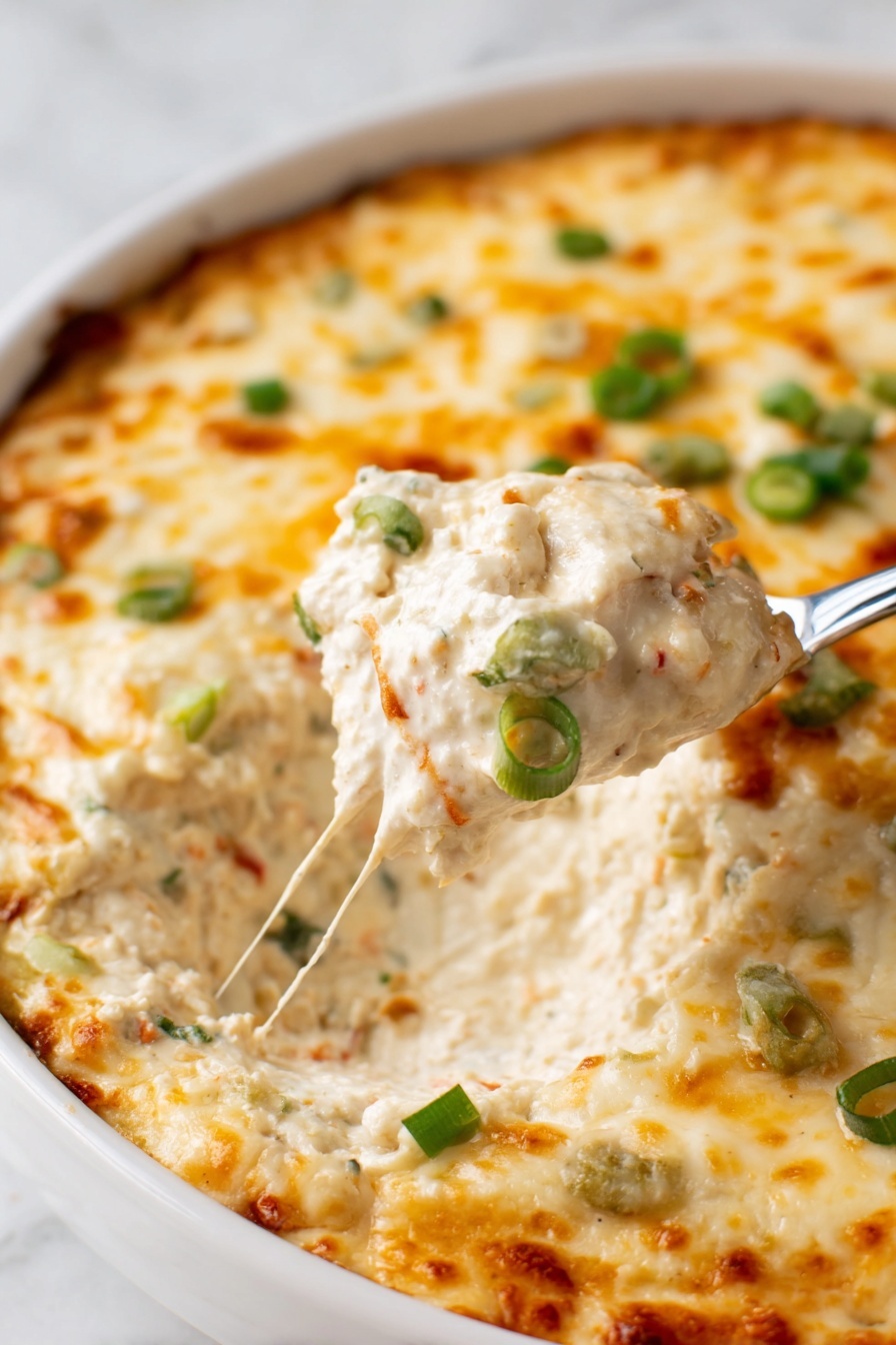 Crab Rangoon Dip with Wonton Chips, crab dip appetizers, creamy crab dip recipe, easy wonton chip recipe, crowd-pleasing game day dip - A close-up of a white dish filled with a creamy dip that has a thick melted cheese layer on top. The cheese is golden with light brown spots and sprinkled with small green onion pieces. The creamy layer underneath is light beige with visible small bits of other ingredients, showing a rich and creamy texture. A spoon is lifting a scoop of the dip, revealing the thick inside. The dish is on a white marbled surface. photo taken with an iphone --ar 2:3 --v 7