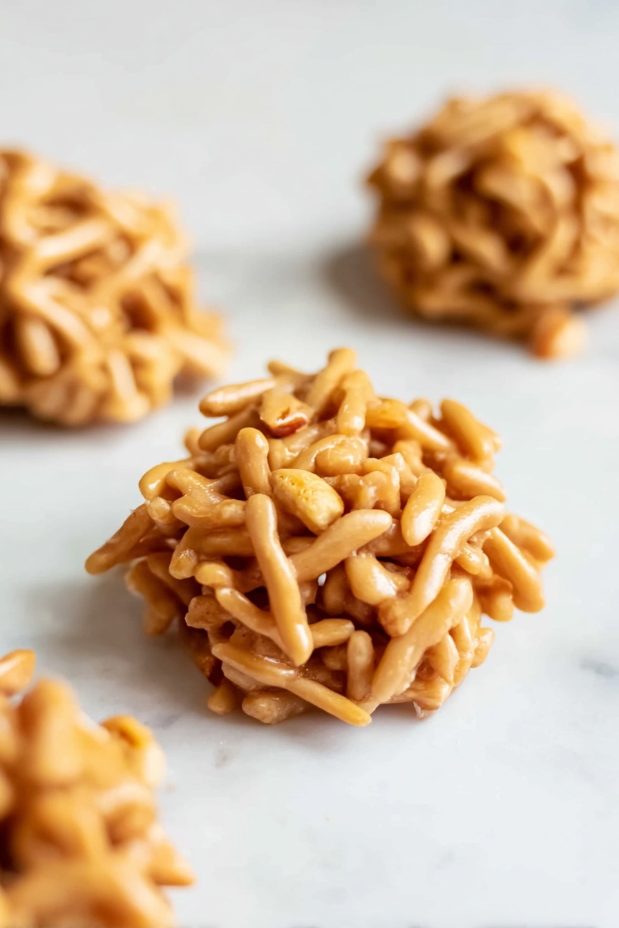 No Bake Peanuts and Chow Mein Cookies, easy no bake cookies, peanut chow mein treats, quick no bake dessert, homemade crunchy cookies - The image shows small round clusters made of light brown, thin, stick-like pieces intertwined with small peanut bits visible in the mix. Each cluster is a single layered mound, with a rough and irregular texture, placed on a smooth white marbled surface. There are three clusters scattered, with one in clear focus at the front and the other two slightly blurred in the background. The sticks have a matte finish with subtle shadows giving depth and softness to the clusters photo taken with an iphone --ar 2:3 --v 7