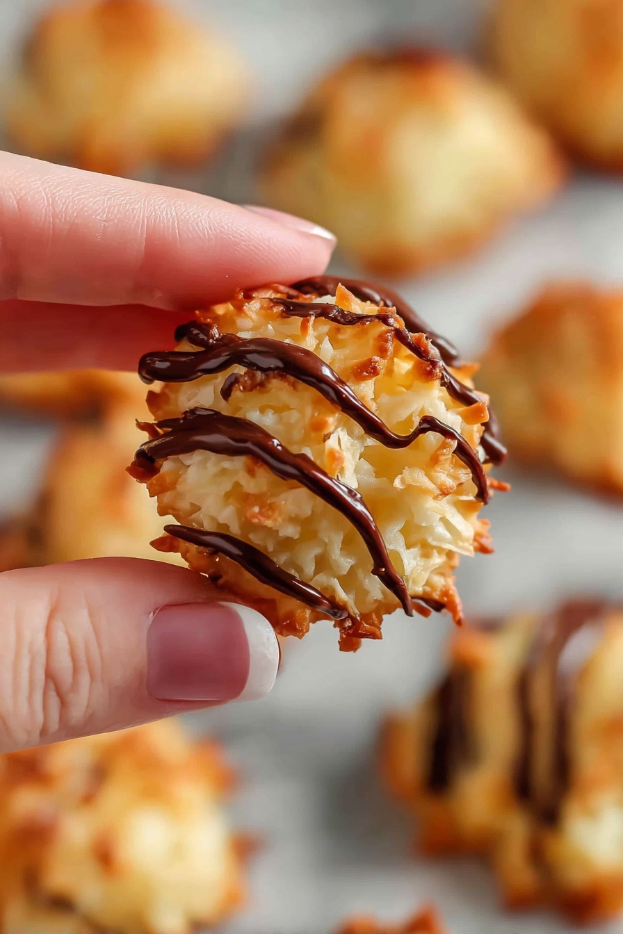 Coconut Macaroons with Chocolate Drizzle, coconut macaroon recipes, easy coconut macaroons, chocolate drizzle desserts, homemade coconut treats - A close-up of a small round cookie made of shredded coconut with a rough, golden-brown surface, topped with several thick dark chocolate drizzles running diagonally across it; the cookie is held delicately between the thumb and woman’s hand index finger, and in the blurry background, more similar cookies are spread out on a white marbled surface. photo taken with an iphone --ar 2:3 --v 7