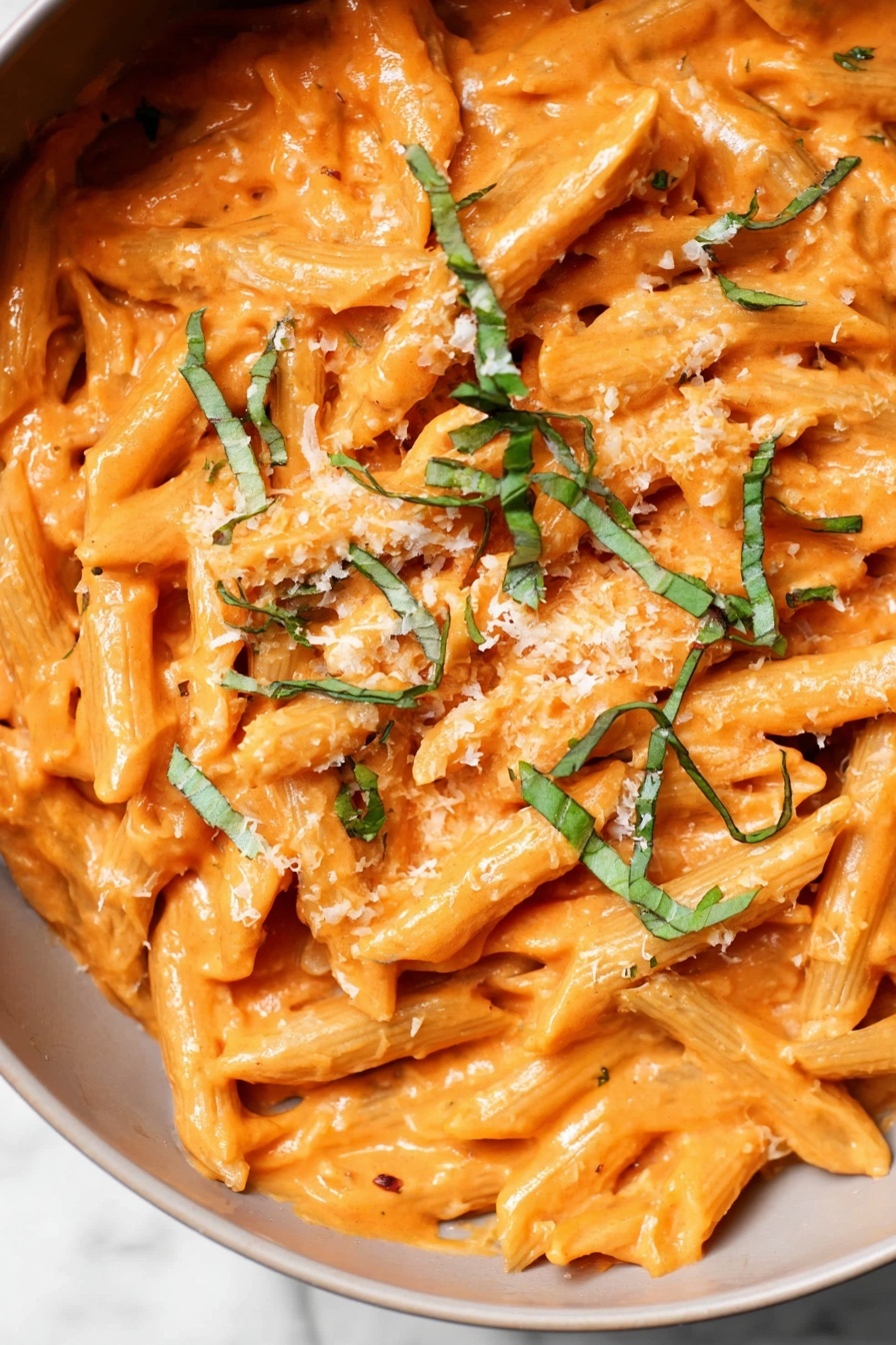 Penne alla Vodka, Penne alla Vodka recipe, creamy pasta, quick pasta dinner, easy vodka pasta - The image shows a close-up of penne pasta covered in a thick, smooth orange sauce. The pasta is mixed well with the sauce and topped with thin green strips of fresh basil scattered across the surface. There is a light sprinkle of grated cheese that adds a slightly grainy texture on top. The background is a white marbled texture, making the colors of the pasta and sauce stand out clearly. photo taken with an iphone --ar 2:3 --v 7