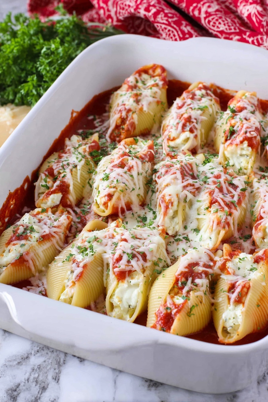 Cheesy Chicken Stuffed Shells, chicken stuffed pasta shells, creamy cheese stuffed shells, easy chicken pasta bake, comforting stuffed shell recipe - The image shows a white baking dish filled with two layers of large pasta shells. The bottom layer is covered with a red tomato sauce, and the shells are stuffed with white cheese inside, visible in some openings. The top layer consists of more pasta shells placed closely next to each other, covered with melted white cheese sprinkled with green herbs and bits of red tomato sauce. The dish is placed on a white marbled surface, with some green herbs and a red patterned cloth in the background. Photo taken with an iphone --ar 2:3 --v 7