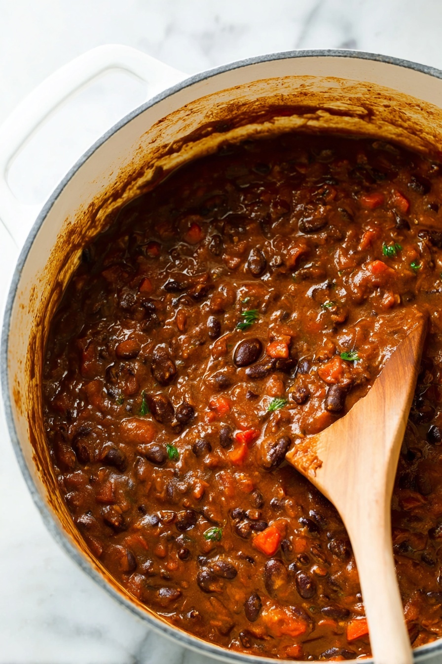 Vegetarian Chili, wholesome vegetarian chili, easy healthy chili, vegan chili recipe, hearty vegetable chili - A close-up view of a thick stew-like dish inside a large white pot with a white marbled surface underneath. The dish has a rich brown color with visible layers of ingredients, including dark brown beans, small red chunks, and some green herb bits mixed throughout. A wooden spoon is partly dipped in the stew on the right side, showing the texture which is soft and slightly chunky with a glossy finish. The sides of the pot have some sauce splashes and the scene looks warm and hearty. Photo taken with an iphone --ar 2:3 --v 7
