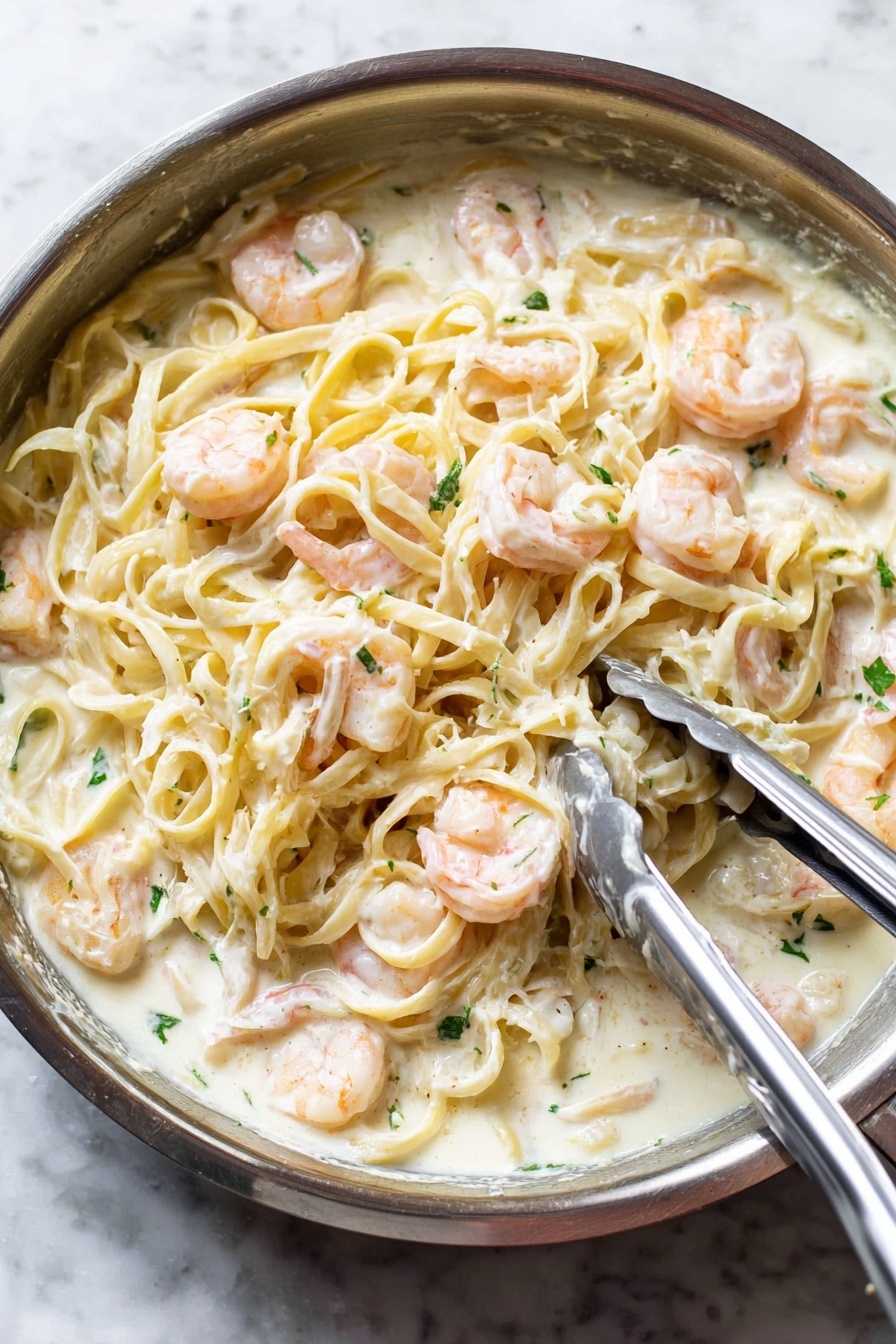 Shrimp Alfredo Pasta, Shrimp Alfredo, Creamy Shrimp Pasta, Easy Shrimp Pasta Recipe, Quick Alfredo Pasta - A close-up view of a silver pan filled with creamy white sauce pasta mixed with pink shrimp. The pasta, light yellow and smooth, is covered in a thick white sauce and evenly spread throughout the pan. Plump shrimp are scattered on top and mixed in, their soft pink color contrasting with the white sauce. Small green herb pieces are sprinkled over the dish for a fresh touch. A pair of metal tongs and a spoon are resting on the pasta, indicating the dish is being stirred or served. The pan sits on a white marbled surface. photo taken with an iphone --ar 2:3 --v 7