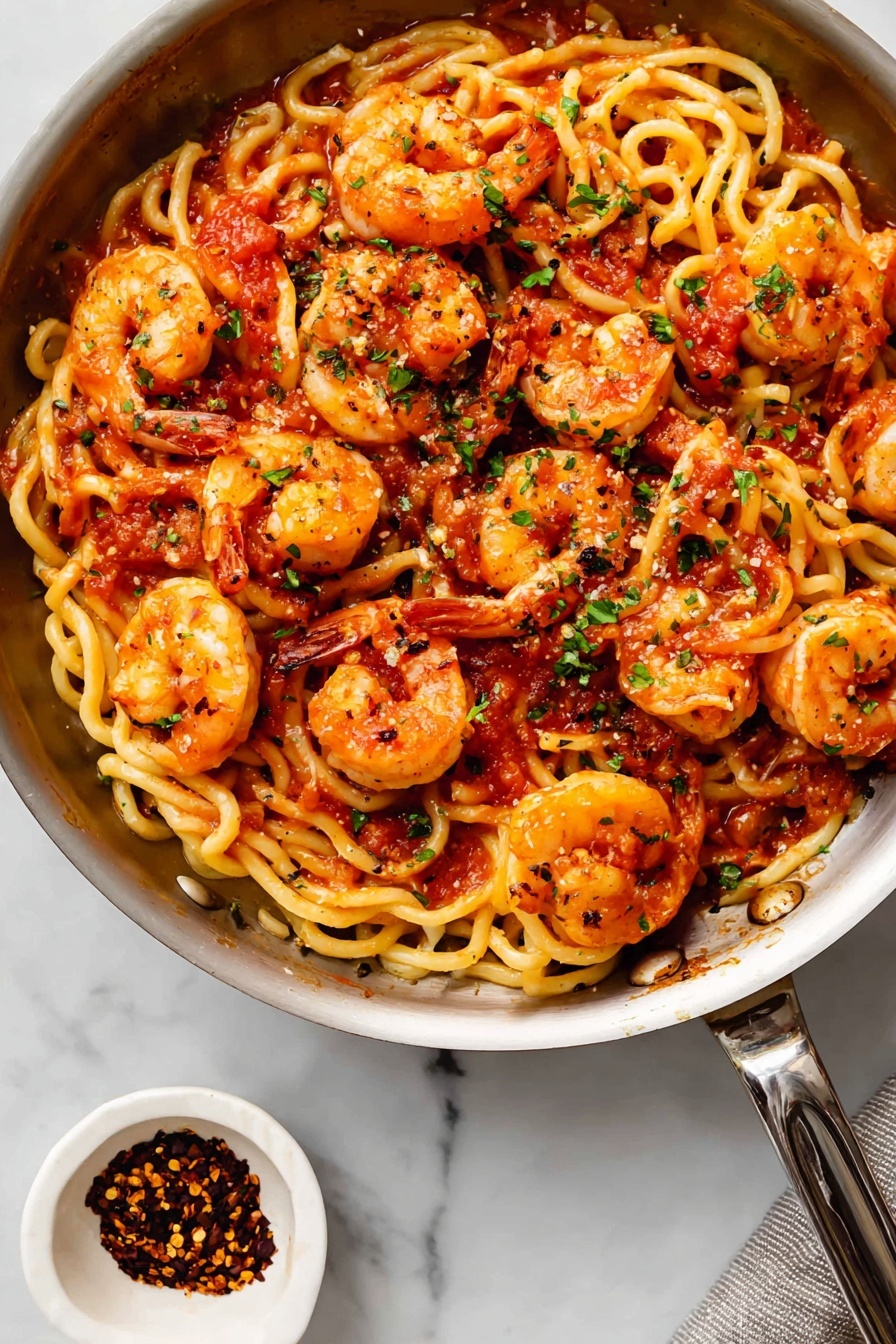 Shrimp Fra Diavolo Pasta, spicy shrimp pasta recipe, garlic shrimp pasta, quick seafood pasta, easy Italian pasta dishes - The image shows a black pan full of curly pasta mixed with cooked shrimp. The pasta is light yellow with a slightly wavy texture, while the shrimp are orange with reddish highlights, laid on top and mixed in all around. There is a red tomato sauce spread unevenly on the pasta and shrimp, and small bits of green herbs sprinkled across the dish. The pan is on a white marbled surface with a small white bowl of red chili flakes beside it at the bottom left. The metal handle of the pan extends out to the right side. Photo taken with an iphone --ar 2:3 --v 7