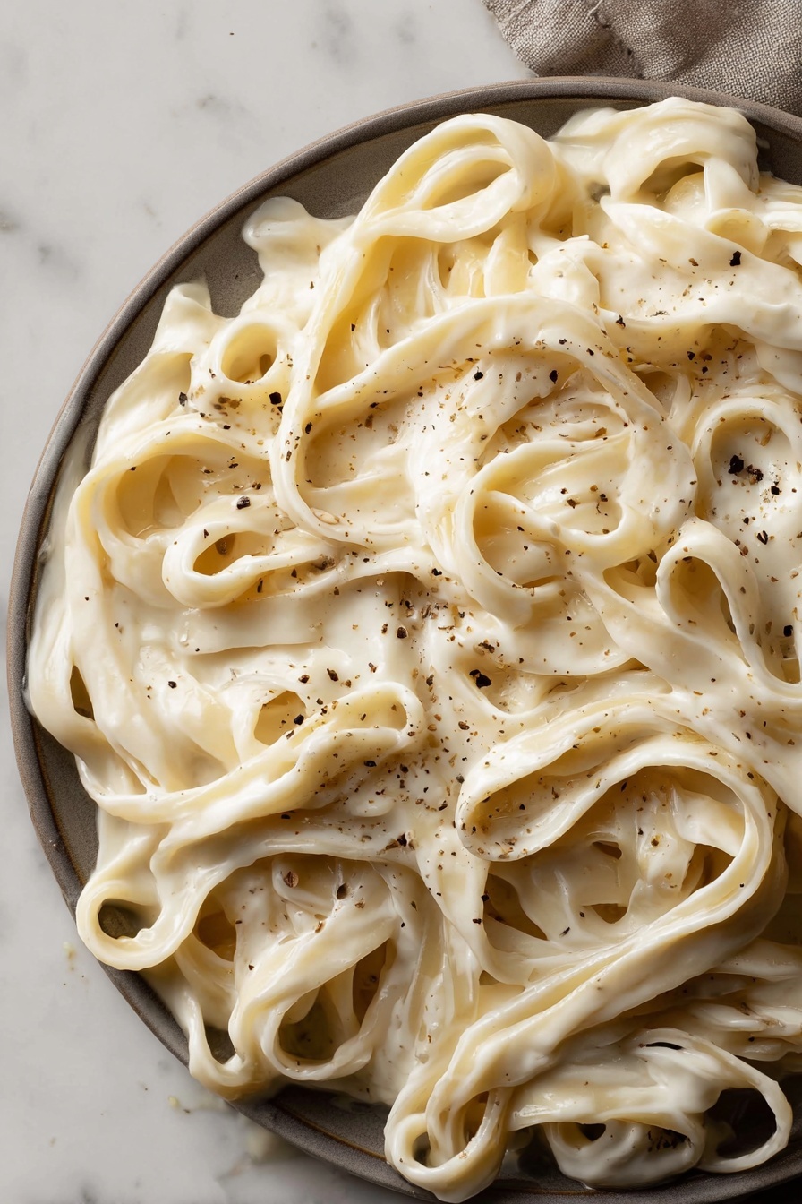 Vegan Cashew Alfredo Sauce, vegan Alfredo sauce, plant-based Alfredo, dairy-free Alfredo sauce, gluten-free Alfredo sauce - This image shows a close-up view of wide fettuccine noodles covered in a thick, creamy white sauce. The sauce looks smooth and rich, evenly coating the noodles. Light brown small specks of black pepper are scattered across the pasta, adding texture and contrast. The dish fills the frame, with noodles tangled and folded over each other in layers. The background is a white marbled surface. photo taken with an iphone --ar 2:3 --v 7