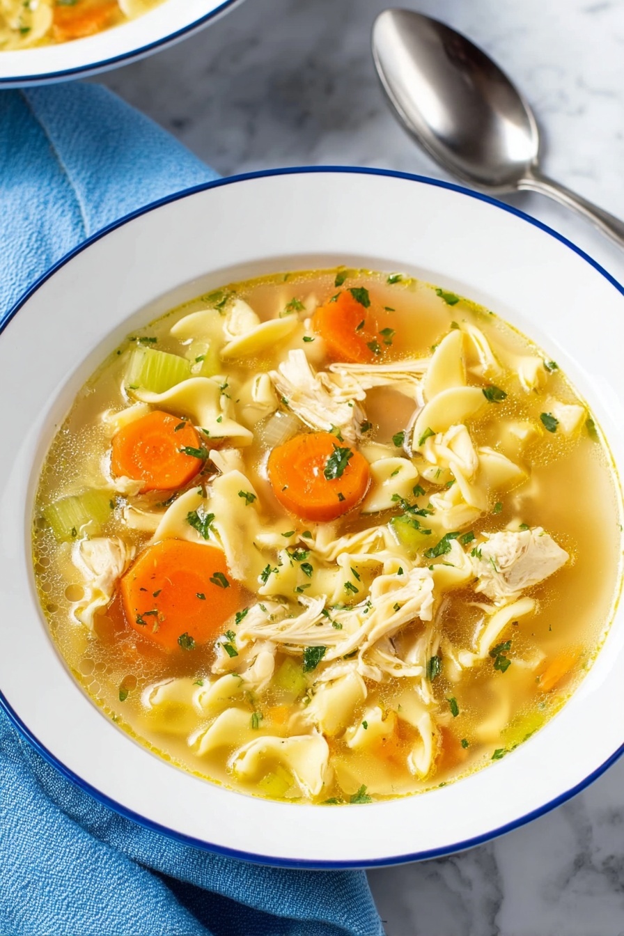 Vegan Chicken Noodle Soup, vegan soup recipe, plant-based chicken noodle soup, healthy vegan soup, comforting vegan noodle soup - A bowl of chicken noodle soup is shown with clear, yellowish broth filling the white bowl with a thin blue rim. The soup has several layers including bright orange carrot slices and pale green celery pieces scattered throughout. Soft, wavy egg noodles are curled in different shapes, mixed with small chunks of cooked chicken that are light beige with a tender texture. Small green parsley flakes are sprinkled on top, adding a fresh touch. The bowl sits on a white marbled surface with a blue cloth and shiny silver spoon nearby. photo taken with an iphone --ar 2:3 --v 7