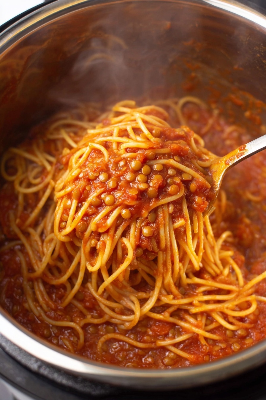 Instant Pot Lentil Spaghetti, healthy instant pot pasta, easy vegetarian pasta recipe, quick lentil dinner, one-pot lentil pasta - A close-up view inside a shiny stainless steel pot filled with spaghetti noodles mixed with small lentils in a rich red tomato sauce; the noodles have a soft yellow color and are coated well by the thick, slightly chunky sauce; a spoon is stirring the mixture, showing a tangled clump of noodles lifted above the rest with pieces of lentils visible around; steam rises gently from the hot dish. photo taken with an iphone --ar 2:3 --v 7