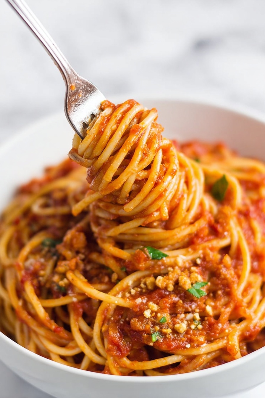 Instant Pot Lentil Spaghetti, healthy instant pot pasta, easy vegetarian pasta recipe, quick lentil dinner, one-pot lentil pasta - A close-up of a white bowl filled with spaghetti noodles coated in rich red tomato sauce with visible small chunks and texture throughout. The spaghetti is thick and twisted around an inserted fork in the center, lifting the noodles upward, showing a dense mix of sauce and soft pasta strands. Small bits of green herbs and crumbled light brown topping are scattered on the surface, adding color contrast. The bowl is placed on a white marbled surface, and the image has a soft background with a focus on the noodles and sauce. photo taken with an iphone --ar 2:3 --v 7