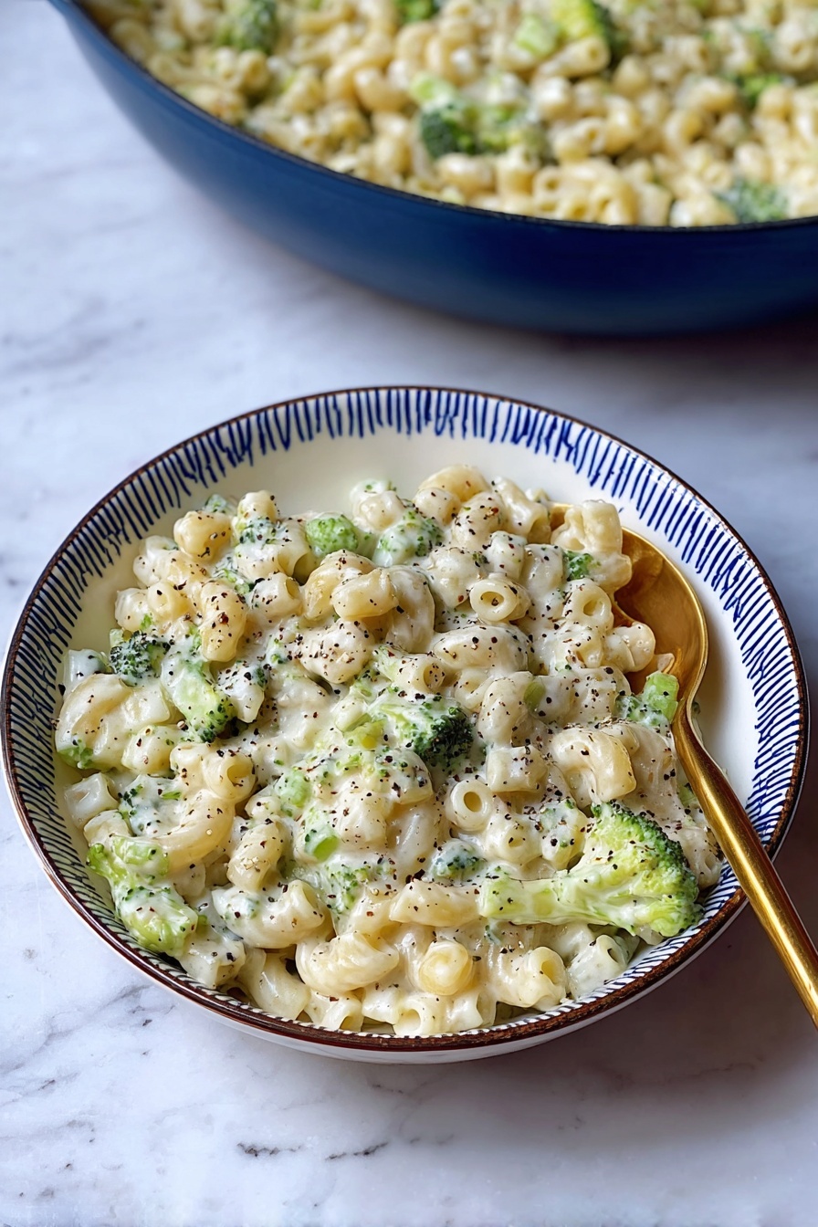 Creamy Broccoli Pasta, creamy broccoli pasta recipe, easy broccoli pasta, vegan broccoli pasta, healthy pasta with broccoli - A white bowl with a blue rim holds a creamy pasta dish made with small tube-shaped pasta mixed with small green broccoli pieces. The pasta is covered in a white, thick sauce, speckled throughout with black pepper. A gold spoon is placed on the right side of the bowl, partially resting in the pasta. Behind the bowl, there is a large blue pan filled with the same creamy pasta and broccoli mixture. The dish is set on a surface with a white marbled texture. photo taken with an iphone --ar 2:3 --v 7