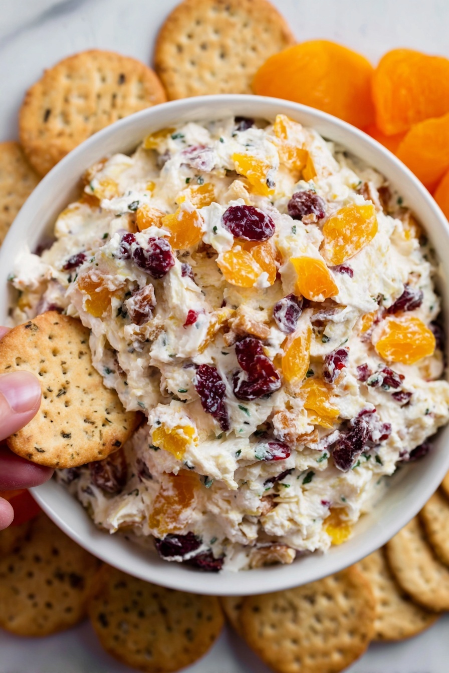 Cranberry Cream Cheese Spread, holiday appetizer, festive cheese spread, easy fruit cheese dip, cranberry cheese appetizer - A white bowl is filled with a creamy mixture that has three layers of visible ingredients: the base is soft white cream cheese, mixed throughout with chunks of orange dried apricots and small dark red dried cranberries, creating a colorful and textured look. Around the bowl, there are beige crackers placed on a white marbled surface, some showing a speckled texture. A woman's hand is holding one of the crackers near the edge of the bowl. The scene is bright and clear, showing the fresh, inviting look of the snack. photo taken with an iphone --ar 2:3 --v 7
