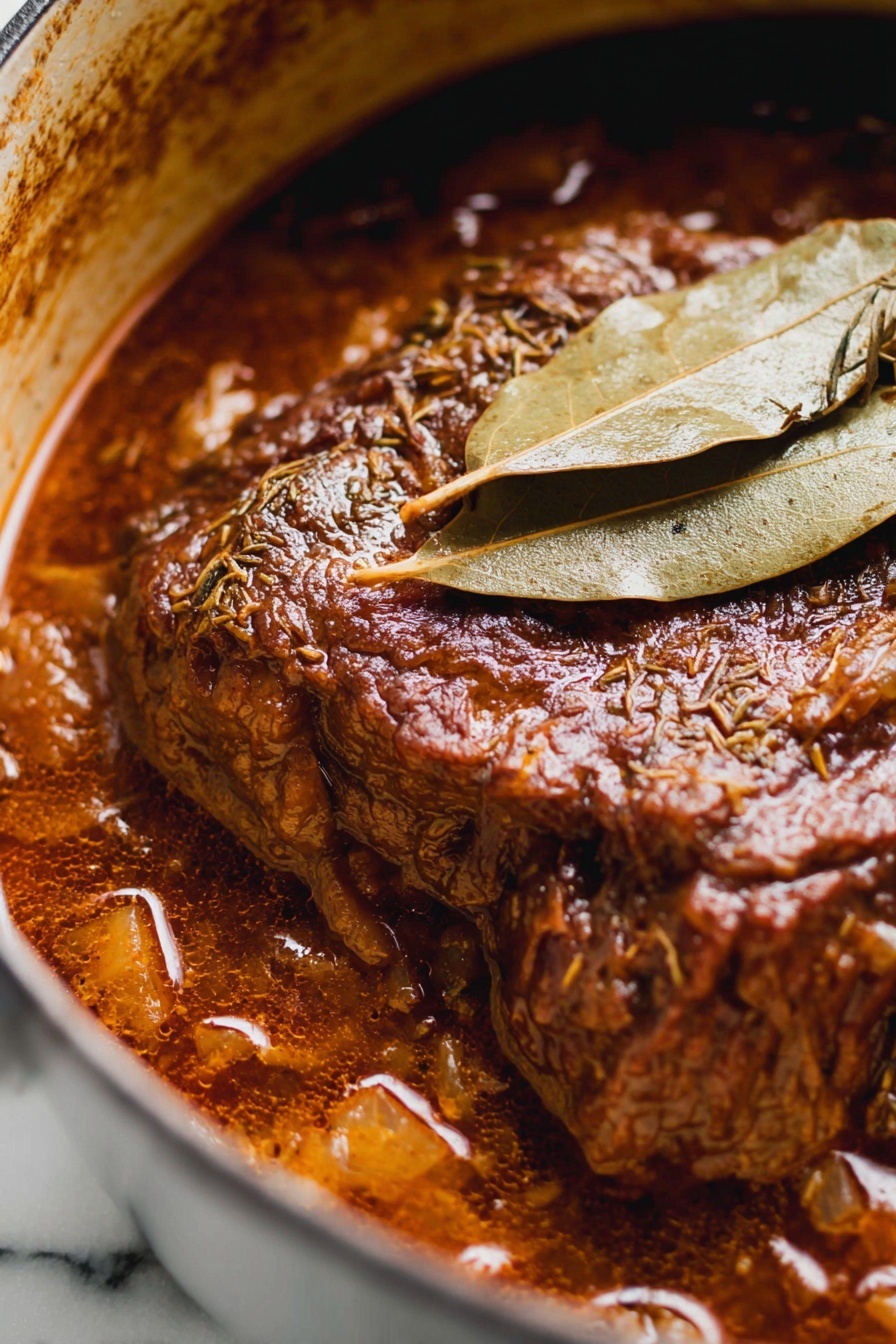 French Onion Beef Pot Roast, French onion soup beef, slow cooker beef roast, hearty beef dinner, caramelized onion roast - The image shows a close-up of a rich, dark brown meat stew in a pot, with the main piece of meat at the center, covered in thick sauce. The meat has a textured surface with visible herbs and spices, and it looks tender and juicy. Two dry, light brown bay leaves rest on top of the meat, adding a layer of texture and color contrast. The stew sauce has a glossy finish with chunks visible around the edges, and the pot’s inner side is stained in a brownish color from the cooking. The background is a white marbled texture. photo taken with an iphone --ar 2:3 --v 7