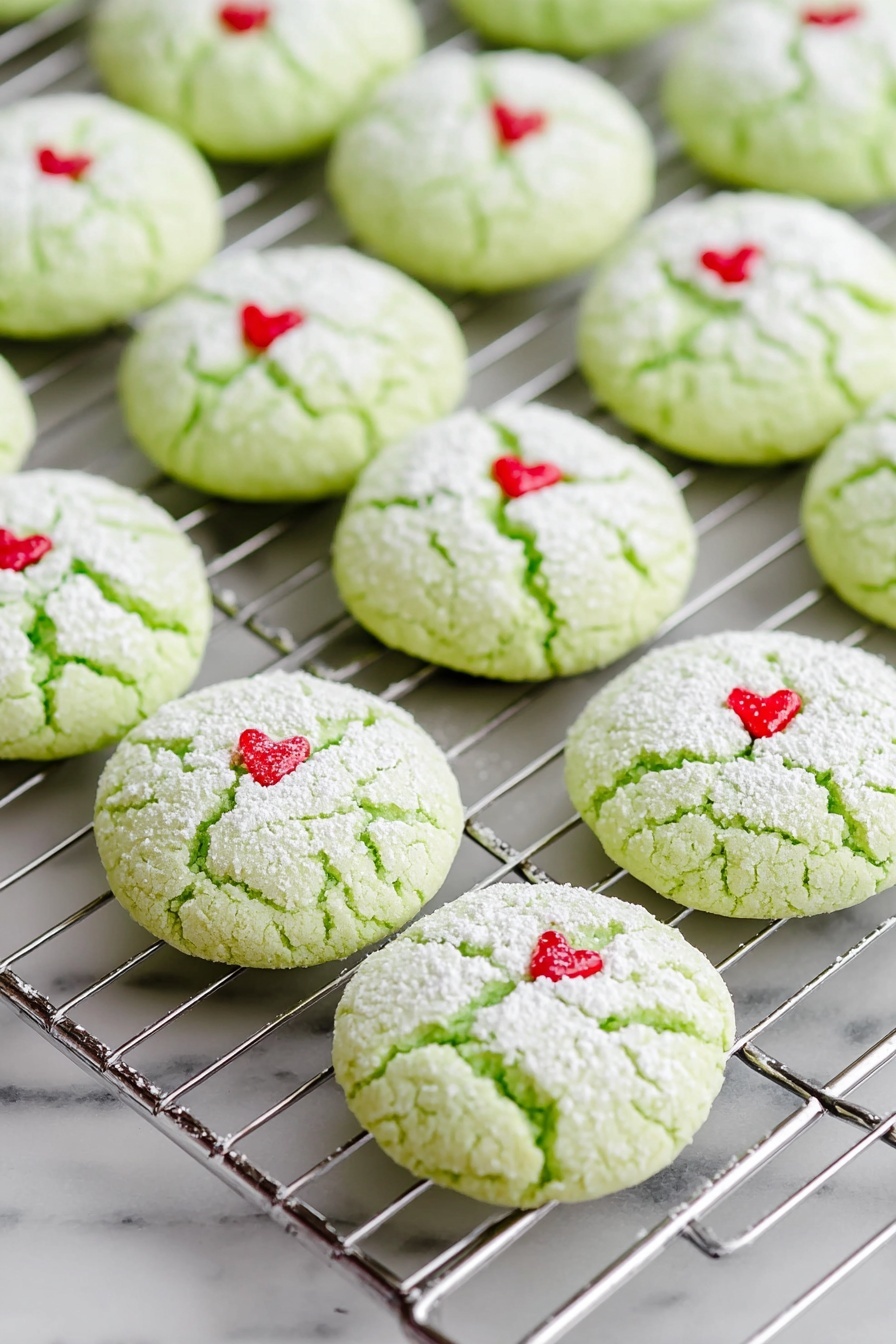 Green Peppermint Cake Mix Cookies, peppermint cake mix cookies, holiday peppermint cookies, easy peppermint cookie recipe, festive green cookies - The image shows rows of light green cookies on a silver cooling rack, each cookie round and cracked on top with a dusting of white powdered sugar. Each cookie has a small red heart-shaped decoration placed near the center. The cooling rack and cookies sit on a white marbled surface that is smooth and clean. The cookies appear soft and slightly puffy with a delicate texture. Photo taken with an iphone --ar 2:3 --v 7