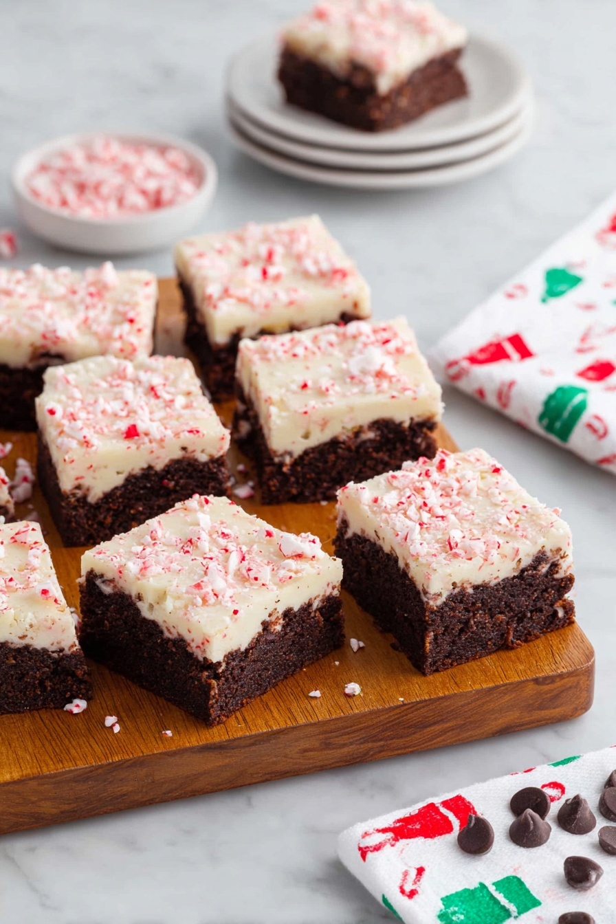 Peppermint Bark Brownies, Peppermint Bark Brownies recipe, holiday brownies with peppermint, festive chocolate brownies, fudgy peppermint brownies - A wooden board holds eleven square brownies, each with two layers: a dark brown, dense textured base with visible chunks inside, topped by a creamy white layer sprinkled with crushed pink peppermint pieces giving a slightly rough texture. Three brownies are separated at the front, showing clear edges and layers. Behind the board, there is a small white stack of four plates with one brownie on top, and to the right, a white napkin with a pattern of small green and red chef hats rests on a white marbled surface. Scattered peppermint candies and a few chocolate chips decorate the bottom edge of the surface. Photo taken with an iphone --ar 2:3 --v 7