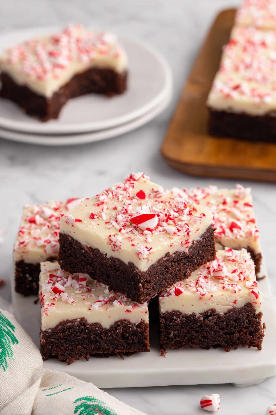 Peppermint Bark Brownies, Peppermint Bark Brownies recipe, holiday brownies with peppermint, festive chocolate brownies, fudgy peppermint brownies - The image shows a stack of four square peppermint brownies on a white plate with a white marbled surface underneath. Each brownie has two layers: a thick, dark brown chocolate base with a moist, dense texture, and a creamy white topping sprinkled with crushed red peppermint bits that add a pop of color and texture. One brownie on top has a small bite taken out, revealing the rich chocolate layer inside. In the background, there is a similar white plate with another brownie on it, and a soft white cloth with green patterns is visible on the side. photo taken with an iphone --ar 2:3 --v 7