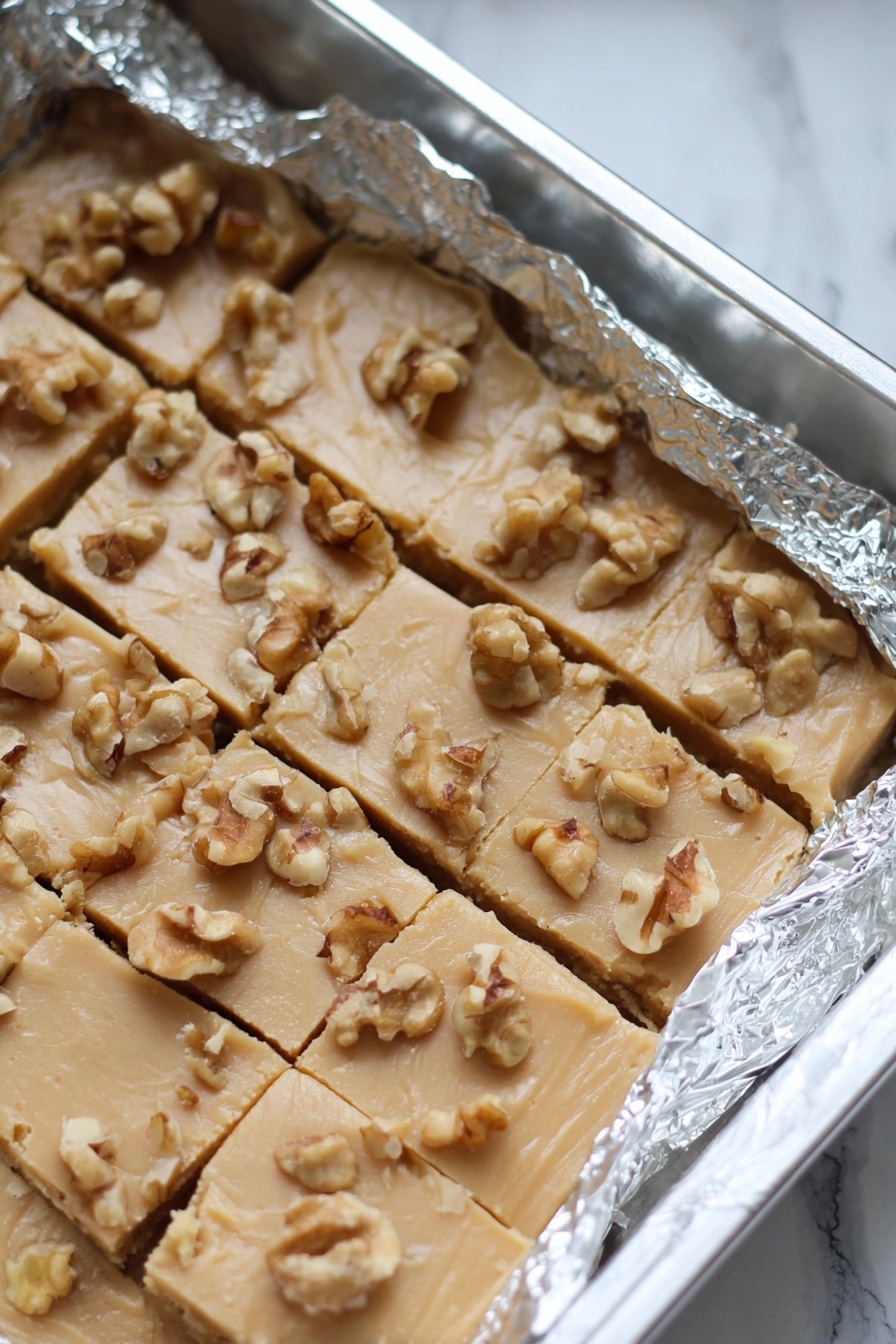 Maple Walnut Fudge, maple walnut fudge recipe, easy maple fudge, homemade walnut fudge, quick fudge with maple and walnuts - The image shows a baking pan lined with crinkled aluminum foil holding a light beige fudge cut into small square pieces, topped with scattered walnut chunks. The fudge has a smooth but slightly cracked texture with the nuts unevenly spread on top. The pan rests on a white marbled surface. photo taken with an iphone --ar 2:3 --v 7