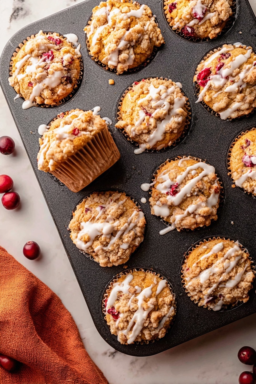 Cranberry Orange Muffins with Crumb Topping, cranberry orange muffin recipe, easy cranberry muffin recipe, moist cranberry muffins, citrus cranberry muffin ideas - The image shows a dark muffin tin with nine muffins inside it, all golden brown with a crumbly topping that is light tan and unevenly spread over each muffin. Bright red berry pieces peek through the muffins, adding spots of color. White glaze is drizzled in thin lines over the top of each muffin, contrasting with the crumbly texture. One muffin is pulled slightly out of its spot showing the sides, which have a ridged paper liner of a light brown color. The muffin tin rests on a white marbled surface with a soft orange cloth partially visible in the bottom left corner and a few dark red whole berries scattered near the tin. photo taken with an iphone --ar 2:3 --v 7