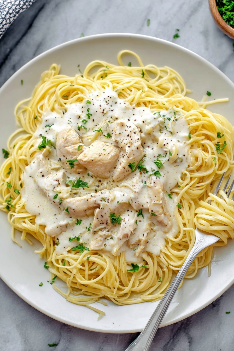 Crock Pot Angel Chicken with Creamy Sauce, crock pot angel chicken, creamy chicken pasta, easy slow cooker chicken, tender chicken with creamy sauce - A white plate holds a dish of thin yellow spaghetti noodles arranged loosely in a circle. On top of the noodles are several pieces of light cream chicken, covered with a thick white creamy sauce that has small bits of herbs and black pepper sprinkled over it. Fresh green parsley leaves are scattered on top of the sauce for a touch of color. A silver fork rests on the right side of the plate, twirling some noodles with sauce. The plate sits on a white marbled surface. Photo taken with an iphone --ar 2:3 --v 7