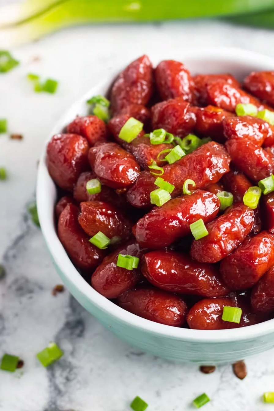 Crockpot BBQ Little Smokies, easiest BBQ appetizer, slow cooker cocktail sausages, crowd-pleasing party snacks, simple game day appetizers - The image shows a white bowl filled with small, shiny red sausage-like pieces covered in a glossy sauce, giving them a sticky and rich appearance. Bright green chopped scallions are scattered evenly over the sausages, adding a fresh contrast in color. The bowl sits on a white marbled surface with a few scattered scallions around it, creating a clean and simple background. The focus is close up and clear, highlighting the texture and color of the sausages and scallions. photo taken with an iphone --ar 2:3 --v 7