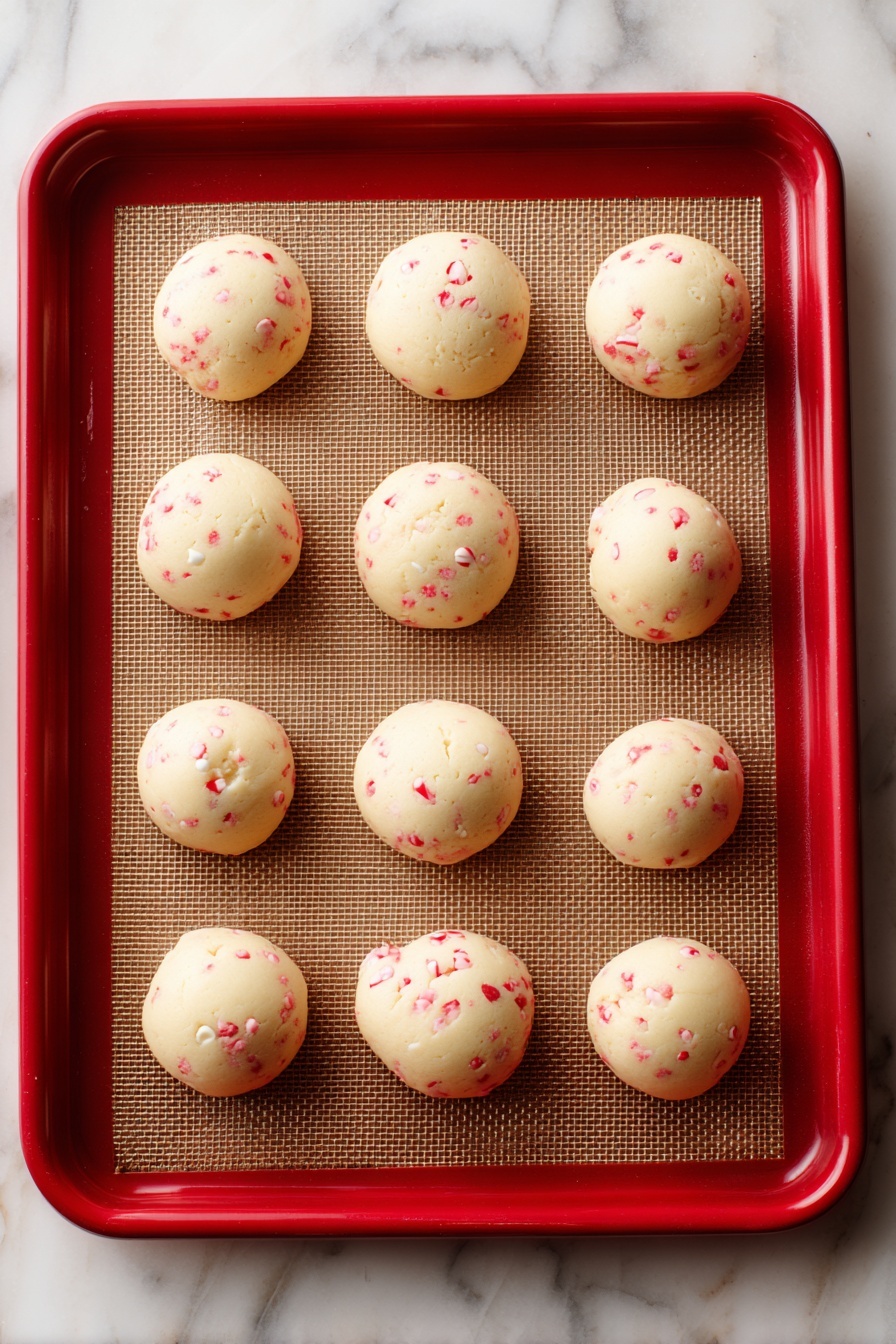 White Chocolate Candy Cane Cookies, holiday peppermint cookies, festive Christmas cookie recipe, easy holiday cookies, peppermint white chocolate treats - The image shows eight round cookie dough balls placed on a brown baking mat lined inside a red-edged tray. Each cookie ball has a creamy, pale dough base with small red specks all over, and some white chocolate chips peek through the dough, adding texture. The surface under the baking mat is a white marbled texture. The dough looks soft, and the balls are evenly spaced apart. photo taken with an iphone --ar 2:3 --v 7