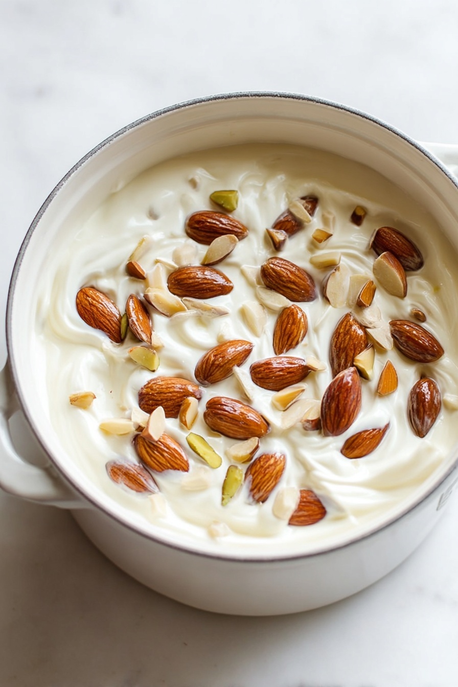 Easy Italian Nougat with Nuts, Italian nougat recipe, homemade nougat with nuts, chewy Italian nougat, traditional Italian torrone - A white pot filled with one main layer of thick, creamy white yogurt mixed with whole and sliced almonds scattered evenly on the top. The almonds vary in shades of light brown to dark brown and are embedded slightly into the creamy yogurt, creating a textured surface with visible swirls in the yogurt beneath. The pot is sitting on a white marbled surface with soft lighting that highlights the smoothness of the yogurt and the natural shine of the nuts. Photo taken with an iphone --ar 2:3 --v 7