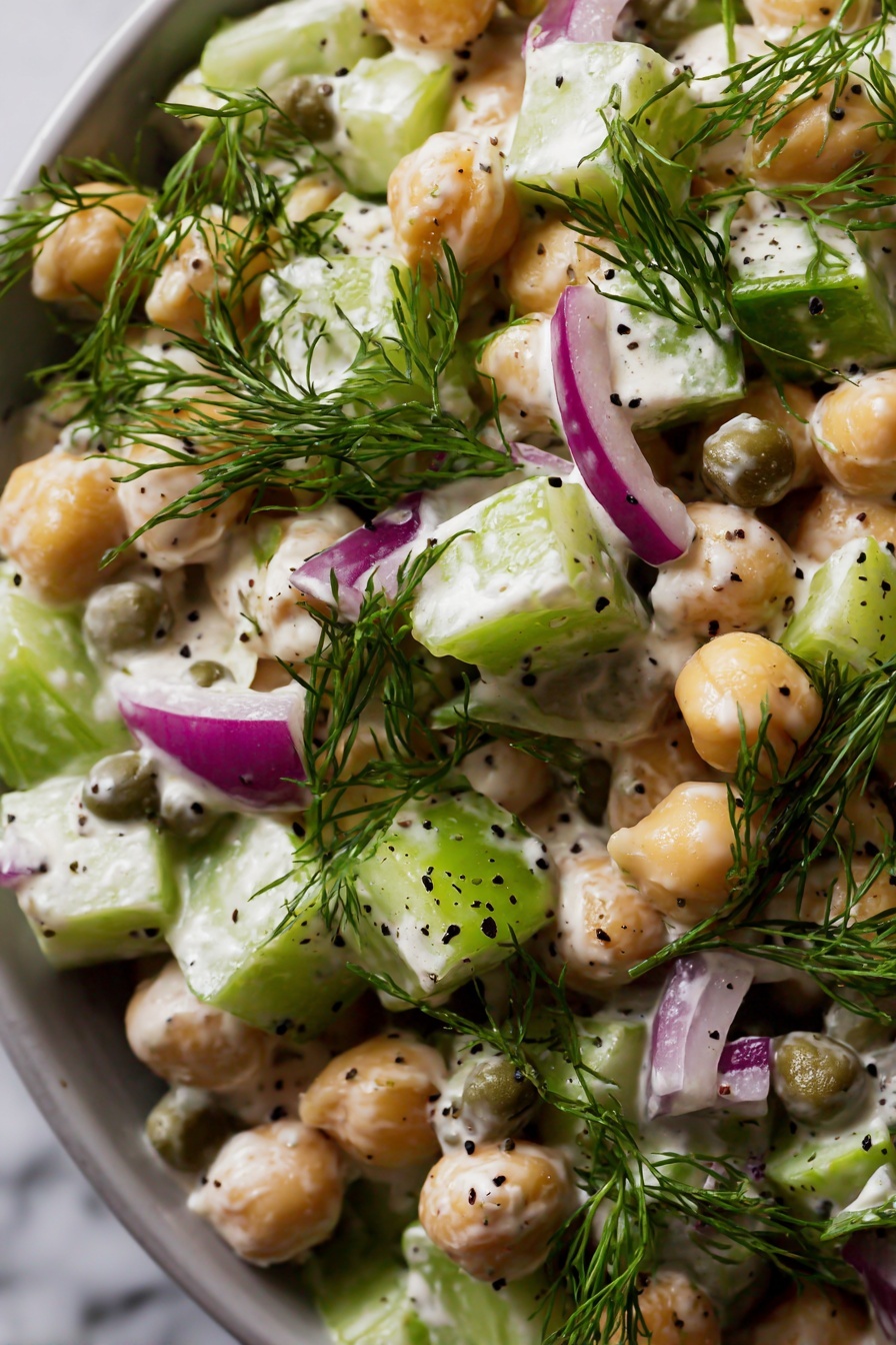 Healthy Tuna Chickpea Salad, healthy tuna salad, chickpea salad recipe, nutritious tuna salad, quick lunch recipes - The image shows a close-up of a salad with several layers and colors. The main elements are round chickpeas that are beige and smooth, mixed with small cubes of light green celery and purplish red onion. There are round, green capers scattered throughout. Fresh green dill sprigs are placed on top, adding a feathery texture. The salad is coated with a creamy, white dressing that has small black seeds mixed in. The entire dish sits against a white marbled surface in soft light. photo taken with an iphone --ar 2:3 --v 7