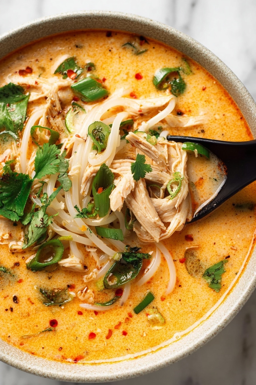 Thai Chicken Curry Soup, Thai Chicken Curry, Easy Thai Soup, Coconut Curry Soup, Spicy Thai Soup - A white bowl filled with creamy orange broth covering soft white noodle strands arranged in the center and slightly tangled. On top, several shredded pale chicken strips are spread out, mixed with small dark green herb pieces and chopped light green onions. A bright green spinach leaf rests near the middle, surrounded by fresh cilantro leaves scattered around the edge. A wedge of lime with visible texture sits on the left side, partially submerged in the broth. The bowl is placed on a white marbled surface. photo taken with an iphone --ar 2:3 --v 7