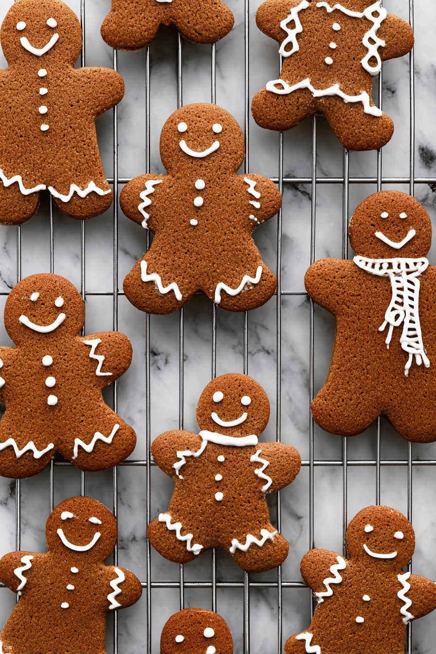 Gingerbread Men Cookies, holiday cookie recipe, baking gingerbread cookies, festive gingerbread treats, homemade gingerbread men - The image shows several gingerbread cookies shaped like people, placed on a metal cooling rack over a white marbled texture. Each cookie is a warm brown color with a rough texture and decorated with white icing in different patterns: some have smiles, eyes, buttons, and zigzag designs on arms and legs, while others have scarves or simple outlines on the edges. The cookies are arranged in various sizes and angles across the cooling rack, creating a cozy and festive feel. photo taken with an iphone --ar 2:3 --v 7