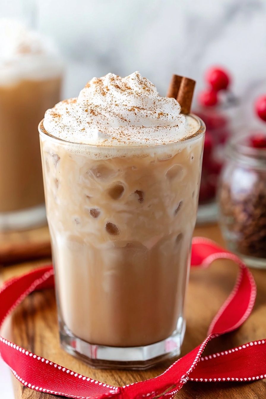 Eggnog White Russian Cocktail, holiday eggnog cocktail, festive White Russian, creamy holiday drinks, Christmas cocktail recipes - A clear glass is filled almost to the top with a creamy light brown drink. On top, there is a tall, fluffy layer of white whipped cream sprinkled with brown spice. A cinnamon stick is placed in the whipped cream on the right side. The glass sits on a wooden surface, with a folded red ribbon with white stitching lying in front of it. There are some small red berries blurred in the background along with other glass containers. The background has a white marbled texture. photo taken with an iphone --ar 2:3 --v 7