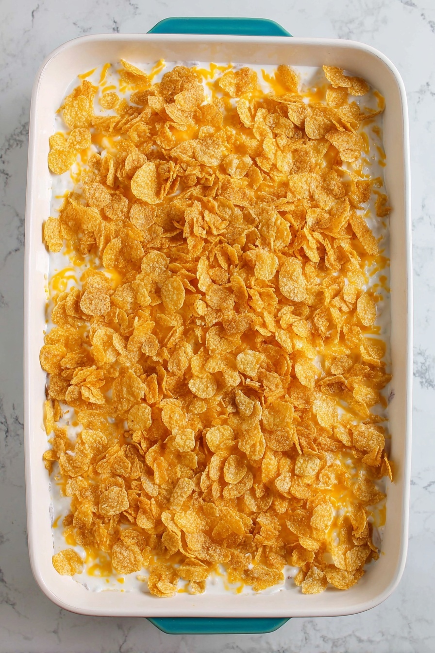 Cheesy Hashbrown Casserole, cheesy hashbrown side dish, cheesy casserole recipe, crispy cheesy potato bake, comfort food hashbrown - The image shows a white rectangular casserole dish filled with a three-layered casserole. The bottom layer is a creamy white sauce with some small visible pieces of ingredients. The middle layer is light orange shredded cheese scattered evenly. The top layer is a generous covering of crunchy golden corn flakes spread all over. The dish is placed on a white marbled surface, and the dish has a teal handle barely visible at the bottom edge. Photo taken with an iphone --ar 2:3 --v 7