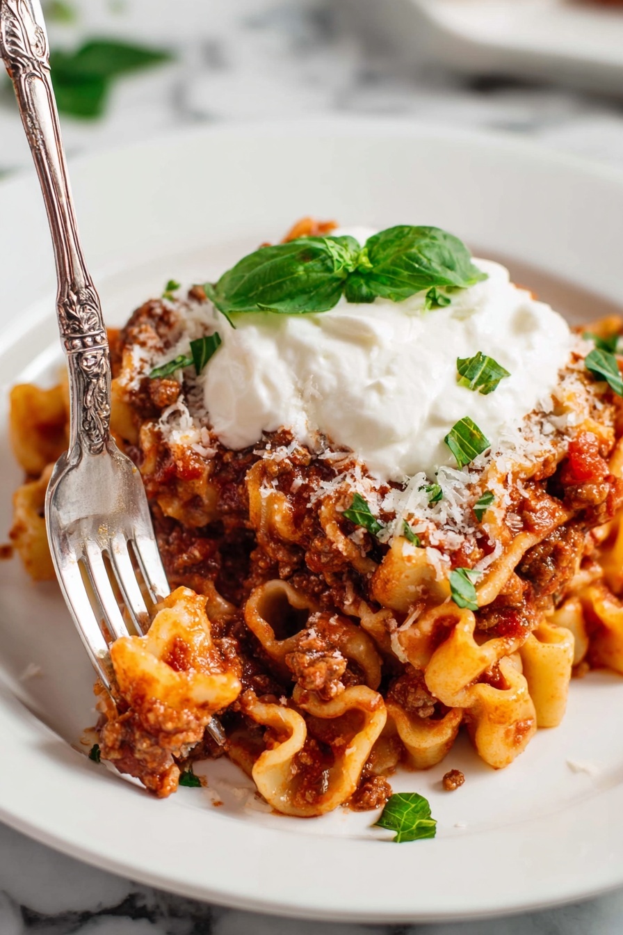 One-Pot Homemade Lasagna Hamburger Helper, easy cheesy lasagna dinner, quick weeknight lasagna, beginner-friendly lasagna recipe, comforting pasta skillet - In the image, there is a white plate on a white marbled surface with pasta mixed with a thick red-brown meat sauce spread across it. On top of the pasta, there is a dollop of white cream with a small green basil leaf placed on it. Thin green basil strips and a sprinkle of shredded white cheese are scattered around the pasta on the plate. A silver fork rests on the edge of the plate. Behind this plate, a larger white bowl filled with more of the pasta dish sits on a light cloth, garnished with a whole basil leaf in the center. To the upper right, part of another white plate with the same pasta dish is visible. Small red cherry tomatoes are scattered at the bottom right corner on the white marbled surface, and a small shaker with red pepper flakes is placed to the left of the main plate. The scene is cozy and inviting, all set on the clean white marble background. photo taken with an iphone --ar 2:3 --v 7