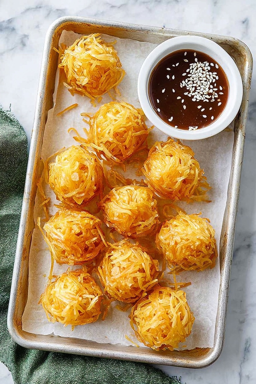 Crispy Shrimp Balls with Spring Roll Coating, Shrimp Balls appetizer, crispy shrimp appetizer, spring roll coated shrimp bites, easy seafood appetizers - The image shows a rectangular metal tray lined with white parchment paper on a white marbled surface. On the tray, there are ten golden-orange crispy balls that look like they are wrapped in thin strips, giving a textured and crunchy appearance. In the top right corner of the tray, there is a small white round bowl filled with a dark brown sauce topped with white sesame seeds, adding a contrast to the crispy balls. A green fabric is partially visible on the left side of the tray. The overall look is neat and appetizing, with the crispy balls clustered but not touching each other, showing off their detailed layers of golden strips. photo taken with an iphone --ar 2:3 --v 7