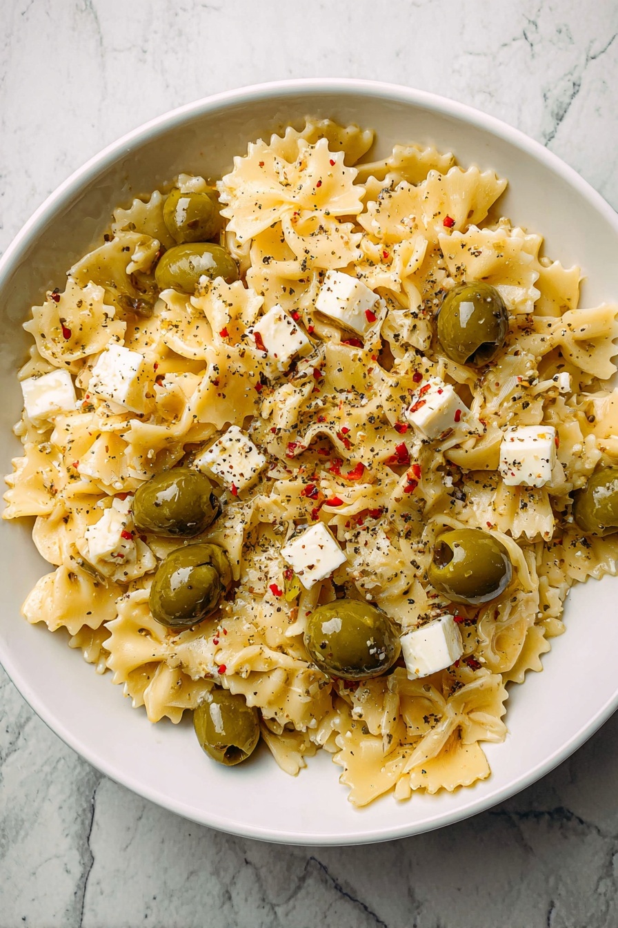 Dirty Martini Pasta Salad, pasta salad with olives and feta, tangy pasta salad recipes, gourmet pasta salad ideas, quick pasta salad dishes - A white bowl filled with pasta made of small, square-shaped pieces with wavy edges, light yellow in color, creating the base layer. Mixed evenly on top are green olives with small pieces of red inside them and small white cubes of cheese scattered throughout. The whole dish is sprinkled with black pepper, adding small dark specks. The bowl is placed on a white marbled surface. photo taken with an iphone --ar 2:3 --v 7