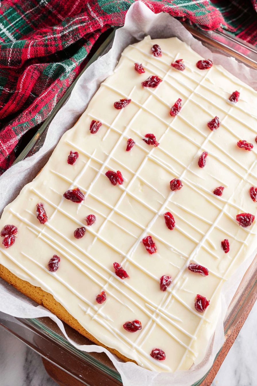 Cranberry White Chocolate Bliss Bars, cranberry white chocolate bars, white chocolate cranberry dessert, holiday cranberry bars, easy white chocolate cranberry recipes - A rectangular layered dessert lies on white parchment paper inside a glass baking dish on a white marbled surface. The bottom layer is golden brown, thin, and even. On top is a thick, smooth white frosting layer covering the entire surface. Thin white icing lines are drizzled across the frosting, forming a light grid pattern. Scattered over the top are small dried red fruit pieces, evenly spaced, adding bright red spots to the white frosting. The cloth nearby has green, red, and dark colors in a check pattern. photo taken with an iphone --ar 2:3 --v 7