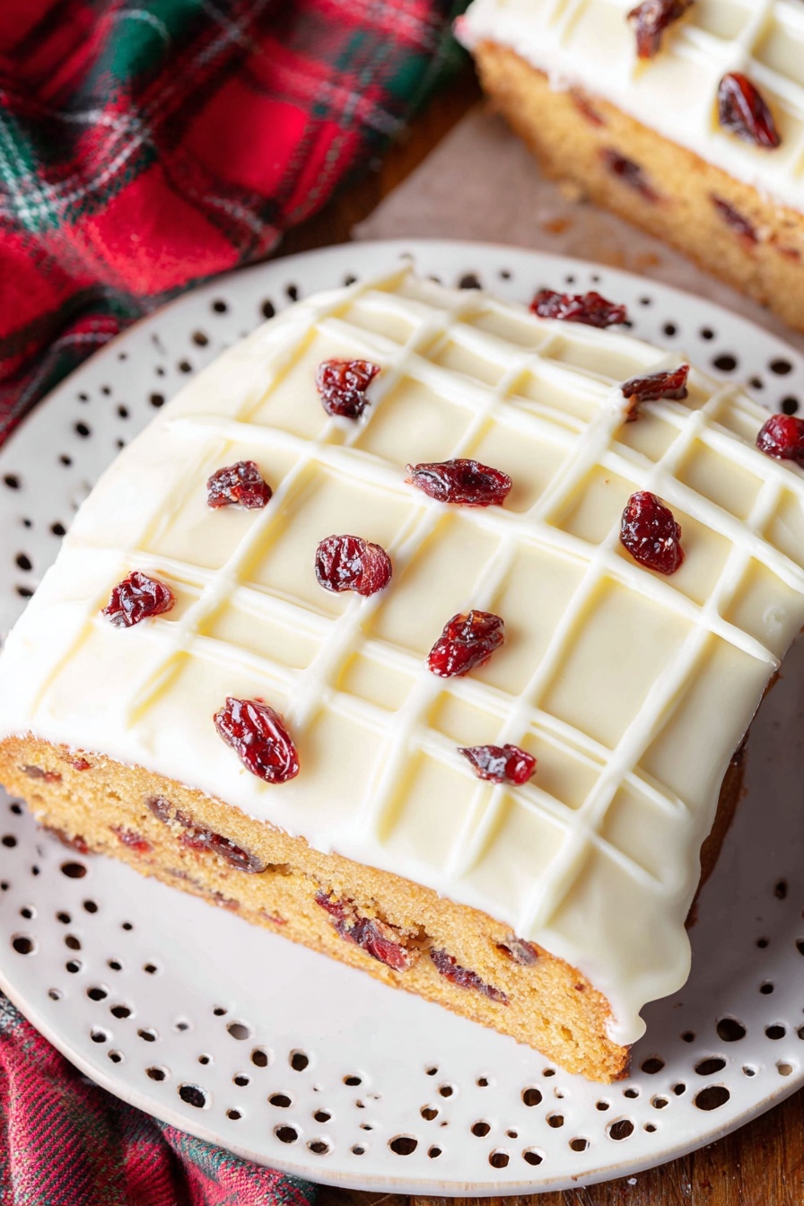 Cranberry White Chocolate Bliss Bars, cranberry white chocolate bars, white chocolate cranberry dessert, holiday cranberry bars, easy white chocolate cranberry recipes - A single slice of cake is shown on a white decorative plate with small cut-out patterns. The cake has two clear layers: a dense golden-brown base with visible small dark red dried fruit pieces inside, and a thick smooth white icing layer on top, decorated with a white grid pattern and scattered dark red dried fruit pieces. The plate rests on a wooden surface with a blurred part of the whole cake and a red and green fabric in the background. Photo taken with an iphone --ar 2:3 --v 7