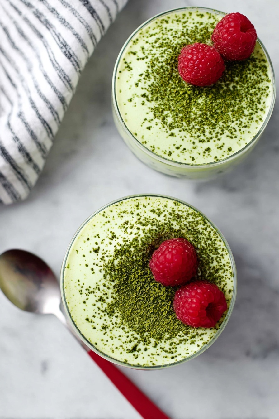 Matcha Protein Chia Pudding, healthy matcha breakfast, vegan chia pudding, high-protein superfood snack, easy overnight chia pudding - Two round glasses filled with a creamy light green mousse topped with a dusting of darker green powder evenly spread on the surface. Each mousse has two bright red raspberries placed near the top edge. The glasses sit on a white marbled surface. A spoon with a red handle is placed next to the bottom glass. Part of a white and black striped cloth is visible on the top left corner. Photo taken with an iphone --ar 2:3 --v 7